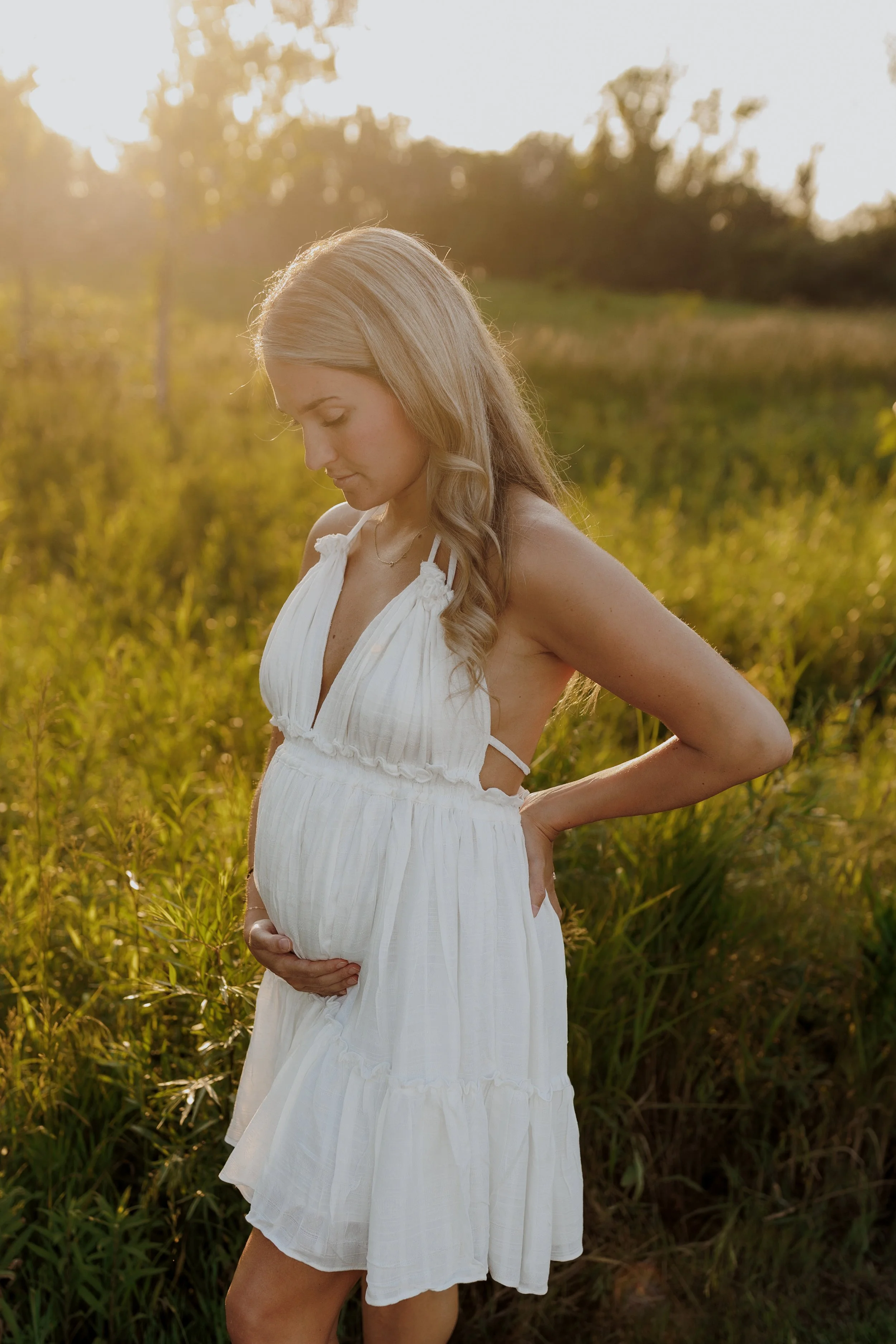A pregnant woman in a white sundress standing in a grassy field during sunset, looking down and touching her belly.
