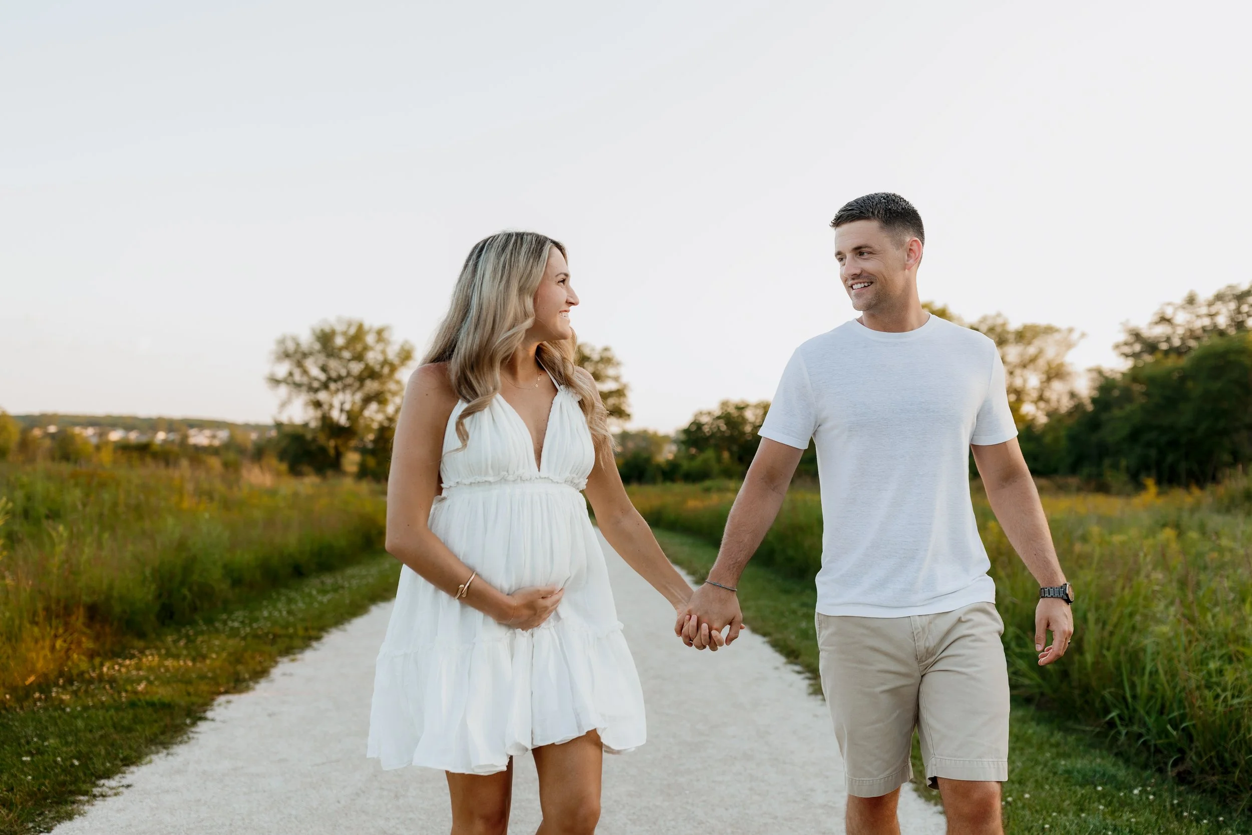 A smiling pregnant woman and a man holding hands while walking outdoors on a dirt path, with trees in the background during the day.