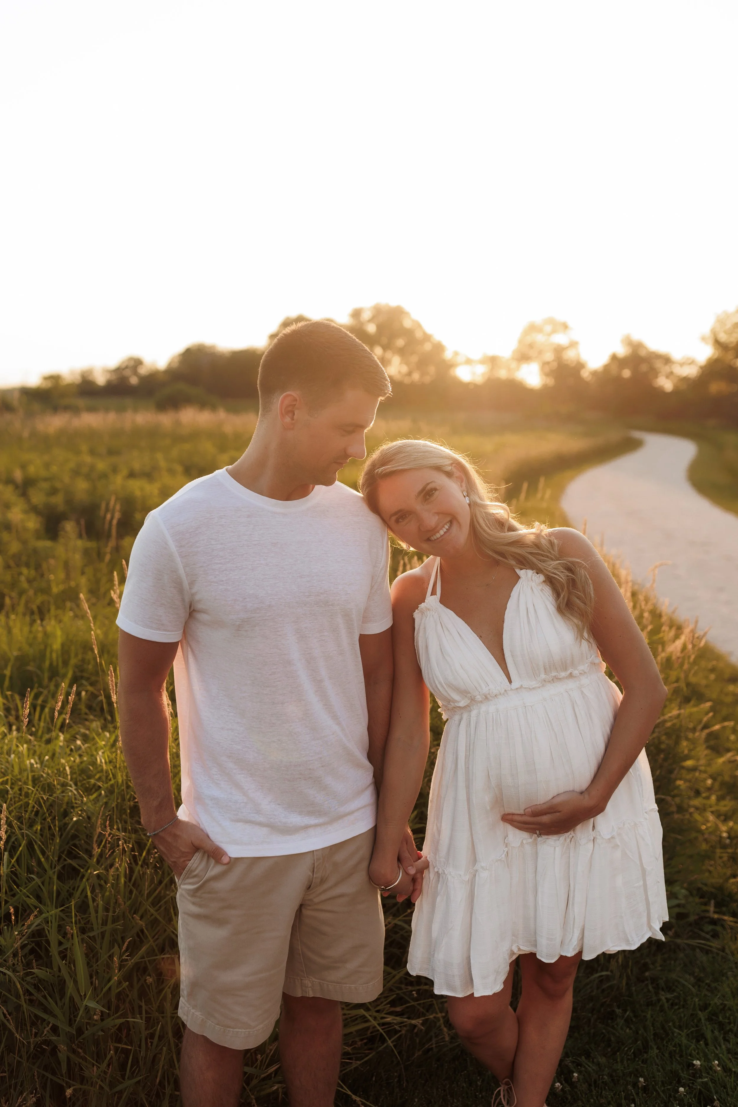 A couple outdoors during sunset, standing close and holding hands, with a woman pregnant, smiling and leaning her head on the man's shoulder, in a grassy field with a winding path and trees in the background.