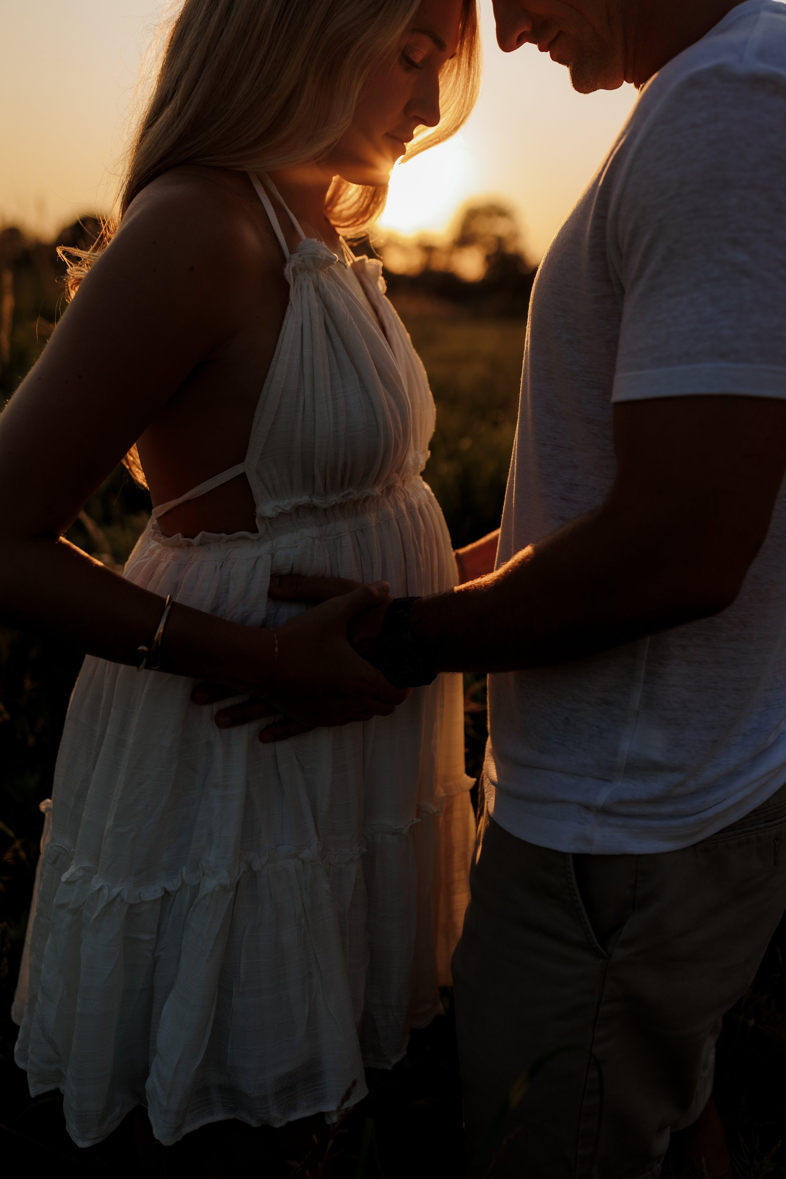 A couple standing close together during sunset, holding hands, with faces close and foreheads touching, in an outdoor setting.