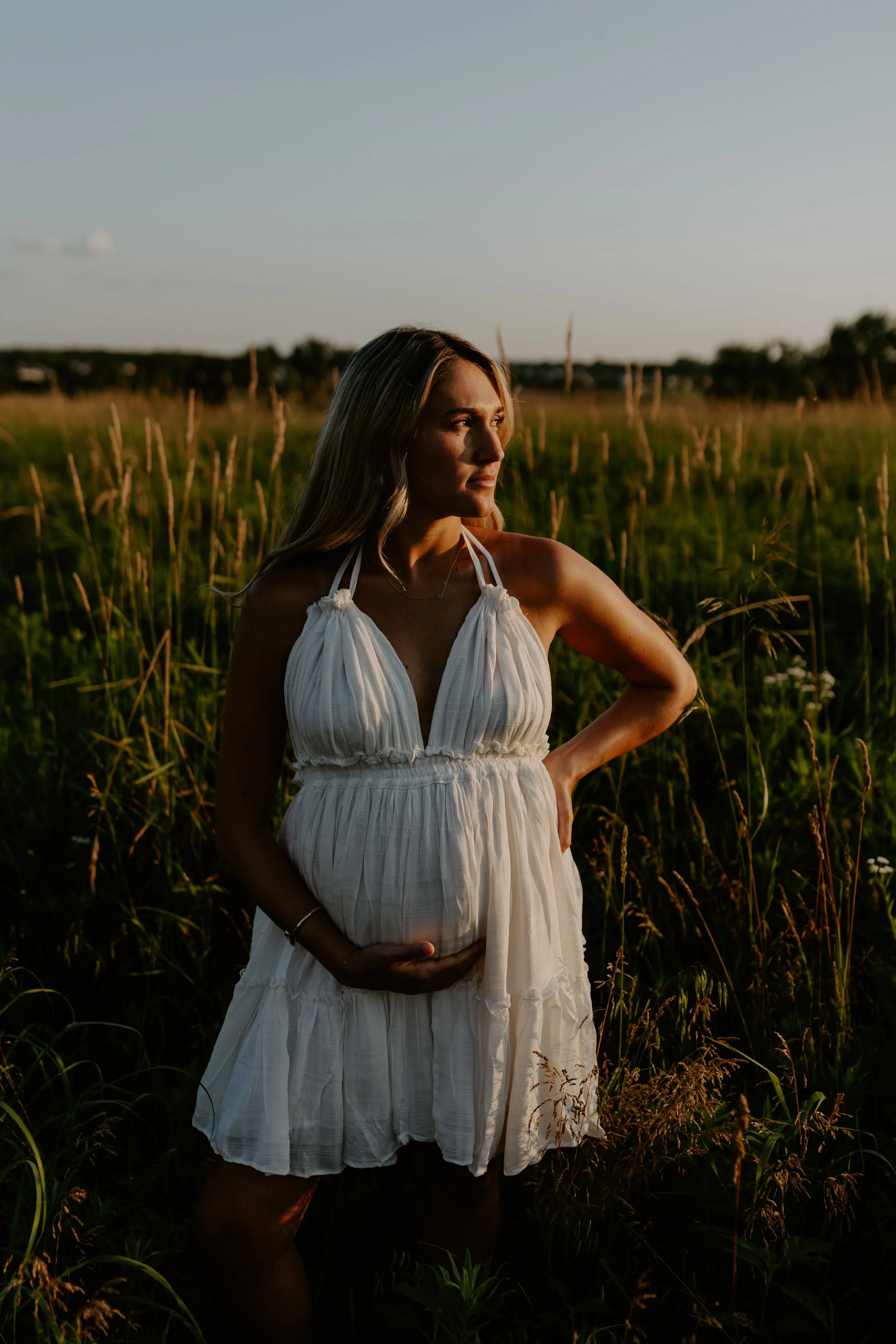 A woman in a white dress standing in a field at sunset with her hand on her stomach and looking to the right.