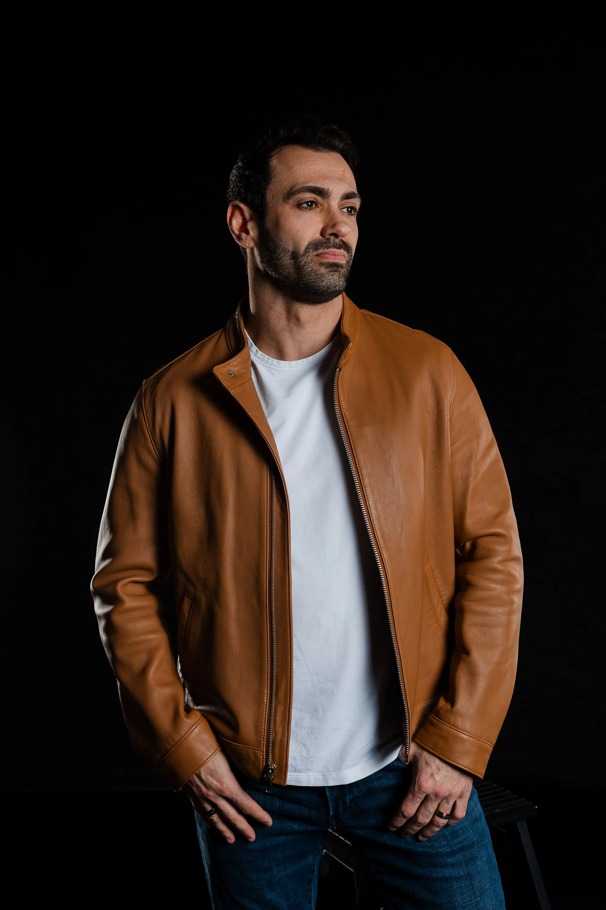 A man with dark hair and a beard wearing a tan leather jacket, white t-shirt, and blue jeans, standing against a black background with a contemplative expression.