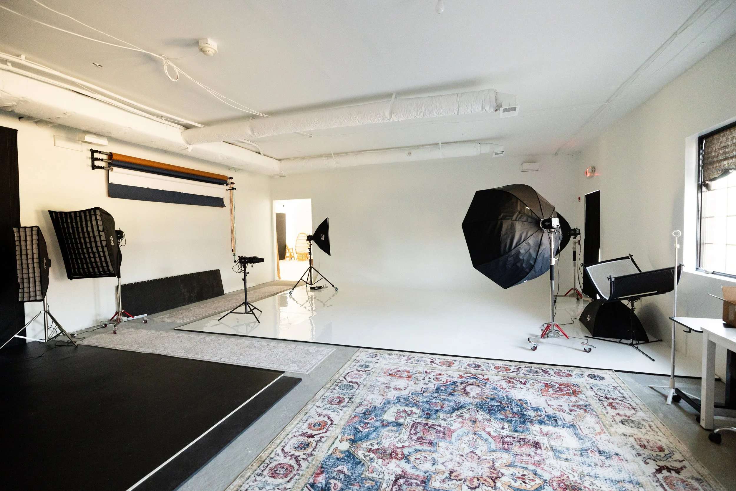 Photography studio with lighting equipment, backdrops, and a white seamless backdrop in a minimalistic room with a large window.