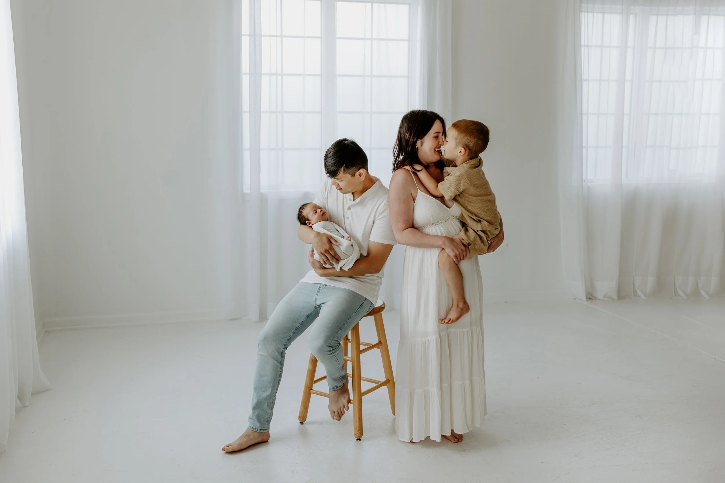 A family of five in a light-filled room with white walls and sheer curtains. A woman and a young boy are standing close, playfully touching noses, smiling, and sharing a joyful moment. A man sitting on a stool holds a newborn baby wrapped in white, w