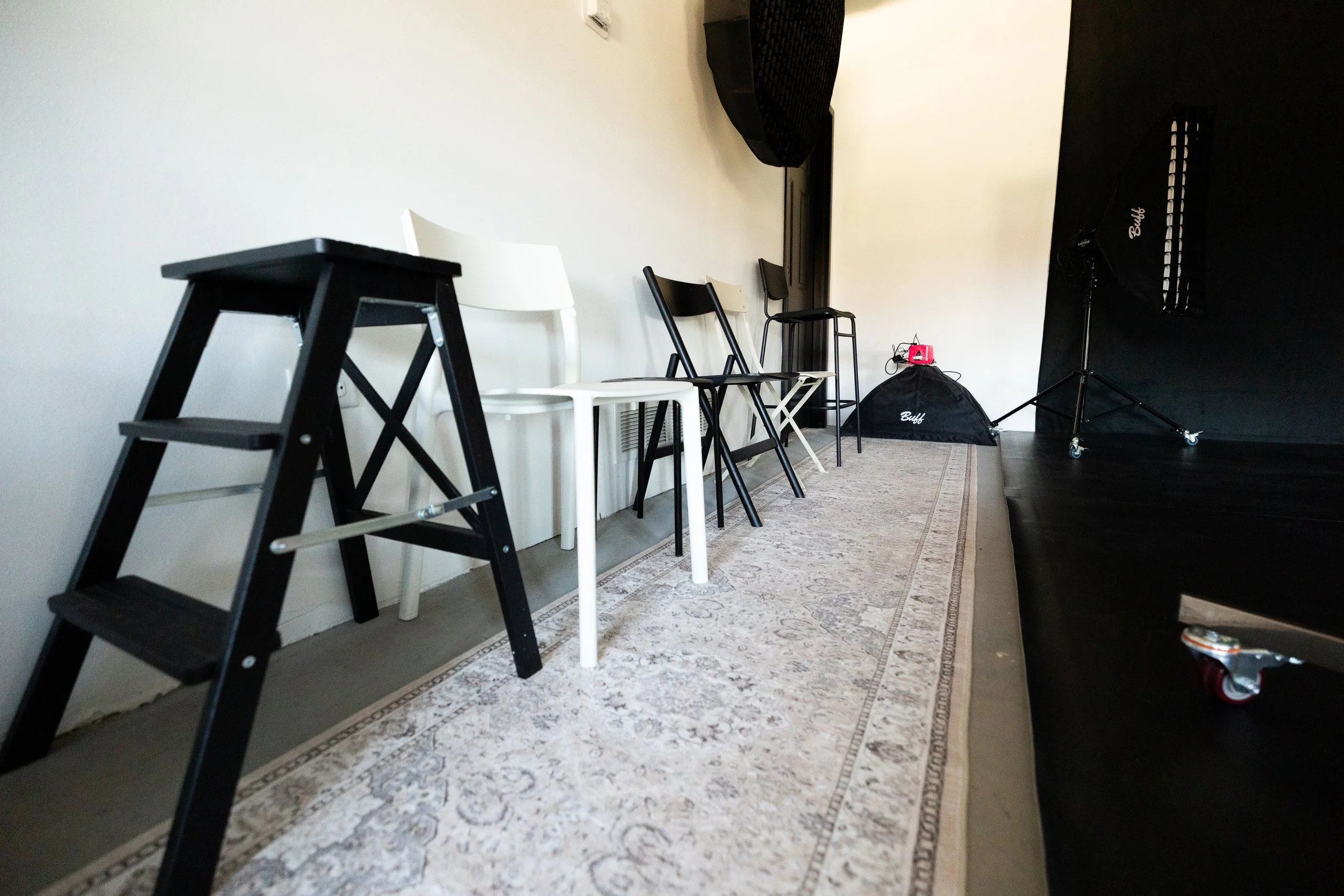 Various chairs in a row on a stage with a white wall and an area rug, including a black step stool, a white chair, black chairs, a black photo backdrop stand with equipment, and a black soft case.