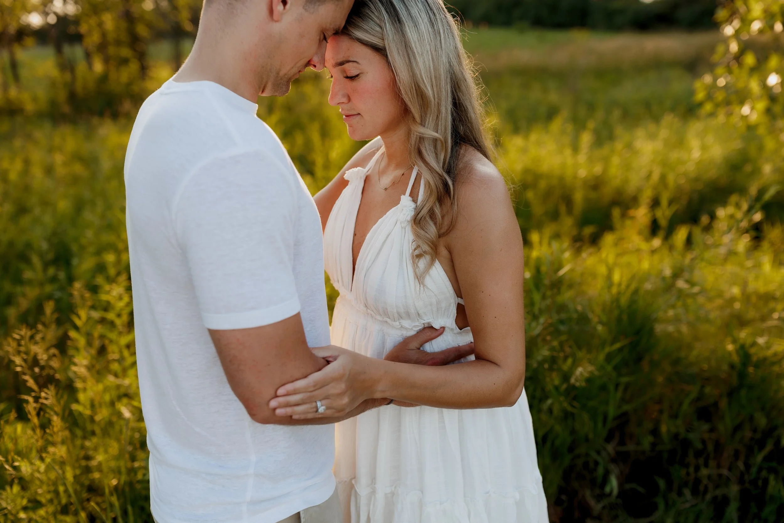 A couple stands close together outdoors during sunset, with foreheads touching and eyes closed, holding hands, dressed casually with the woman in a white dress and the man in a white T-shirt, surrounded by lush greenery.