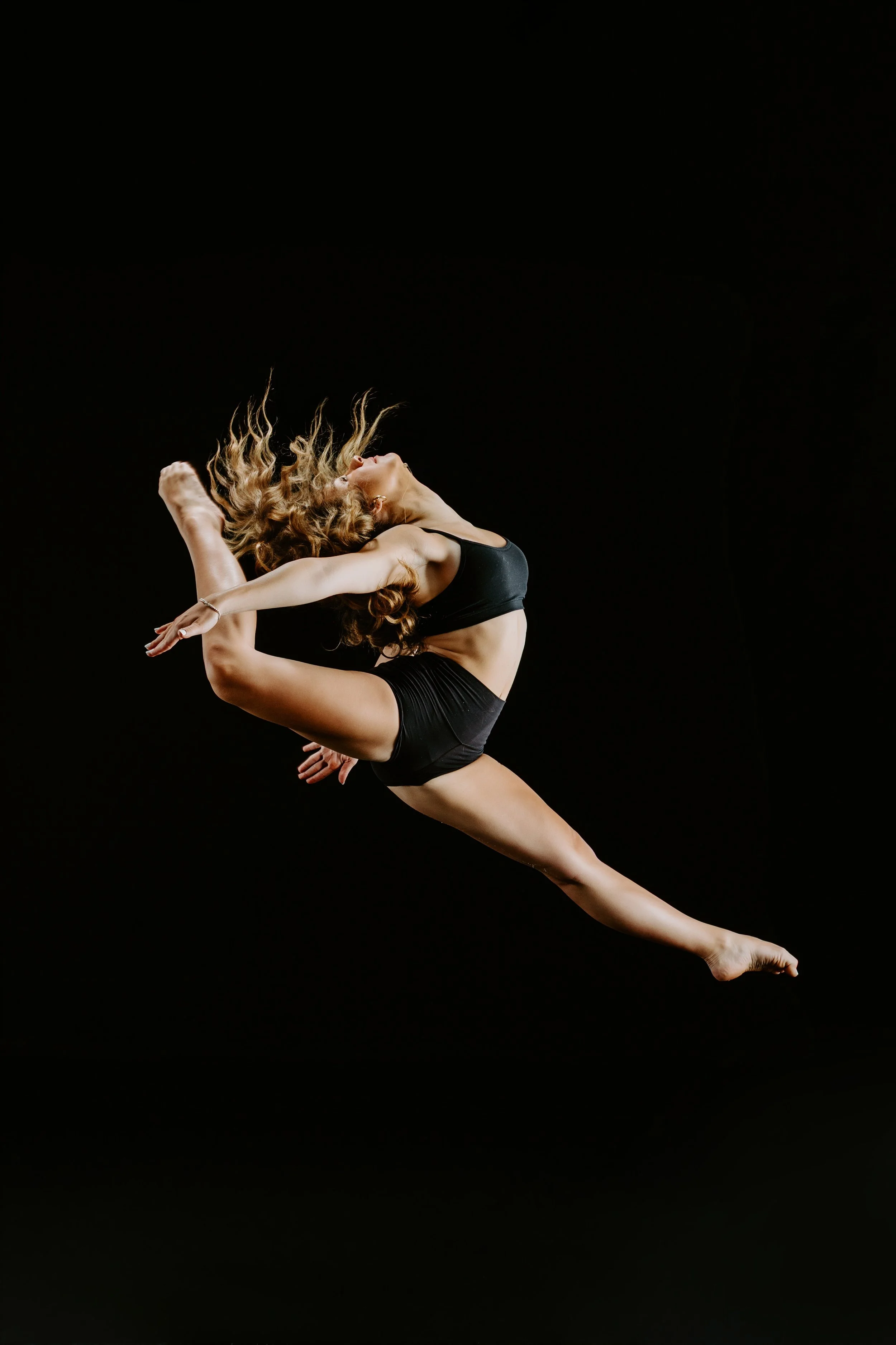 A female dancer performing a leap against a black background, with her body extended and hair flowing.
