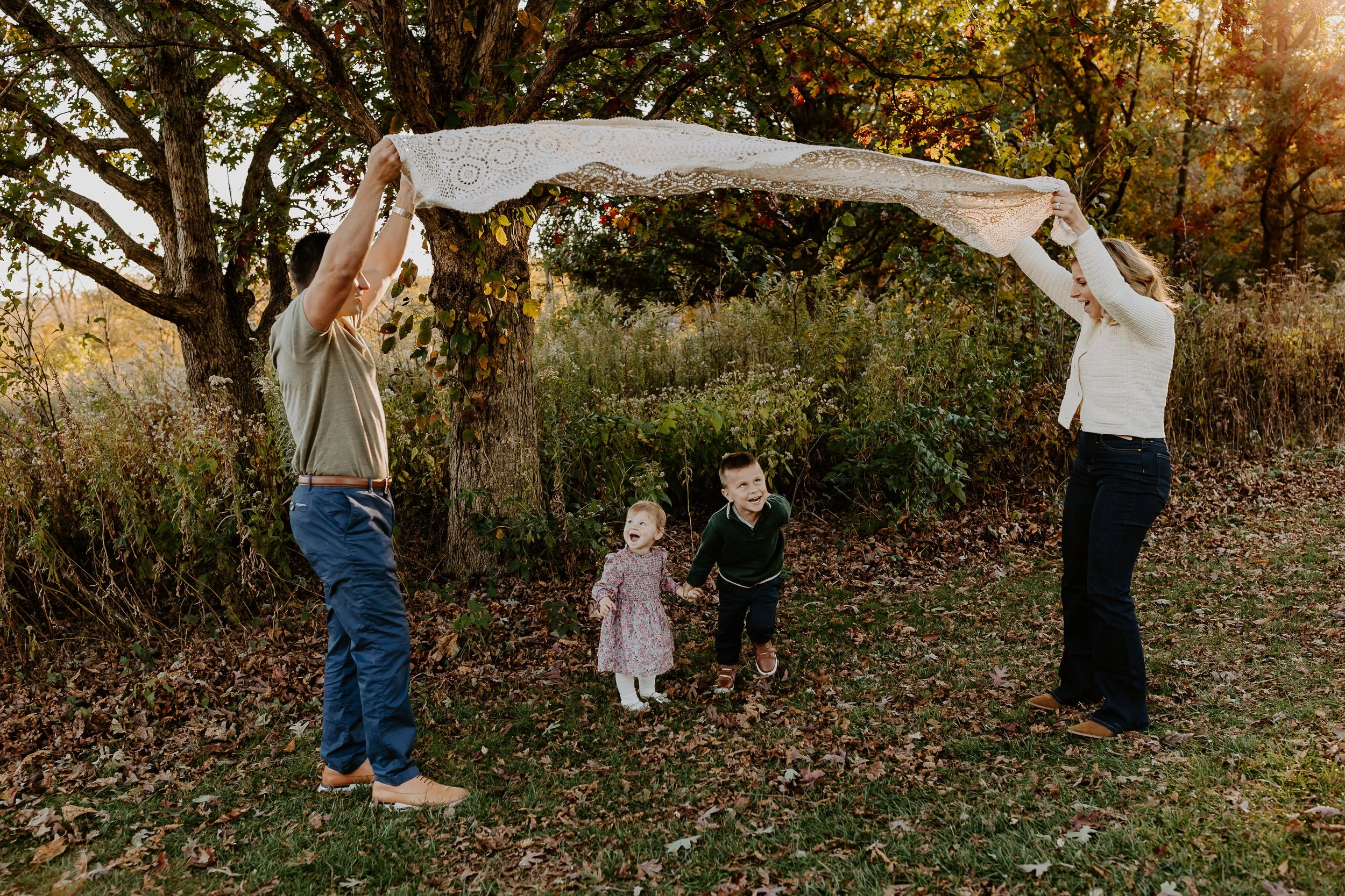 Two women and three children outdoors in a wooded area, holding a lace cloth between them as they play.