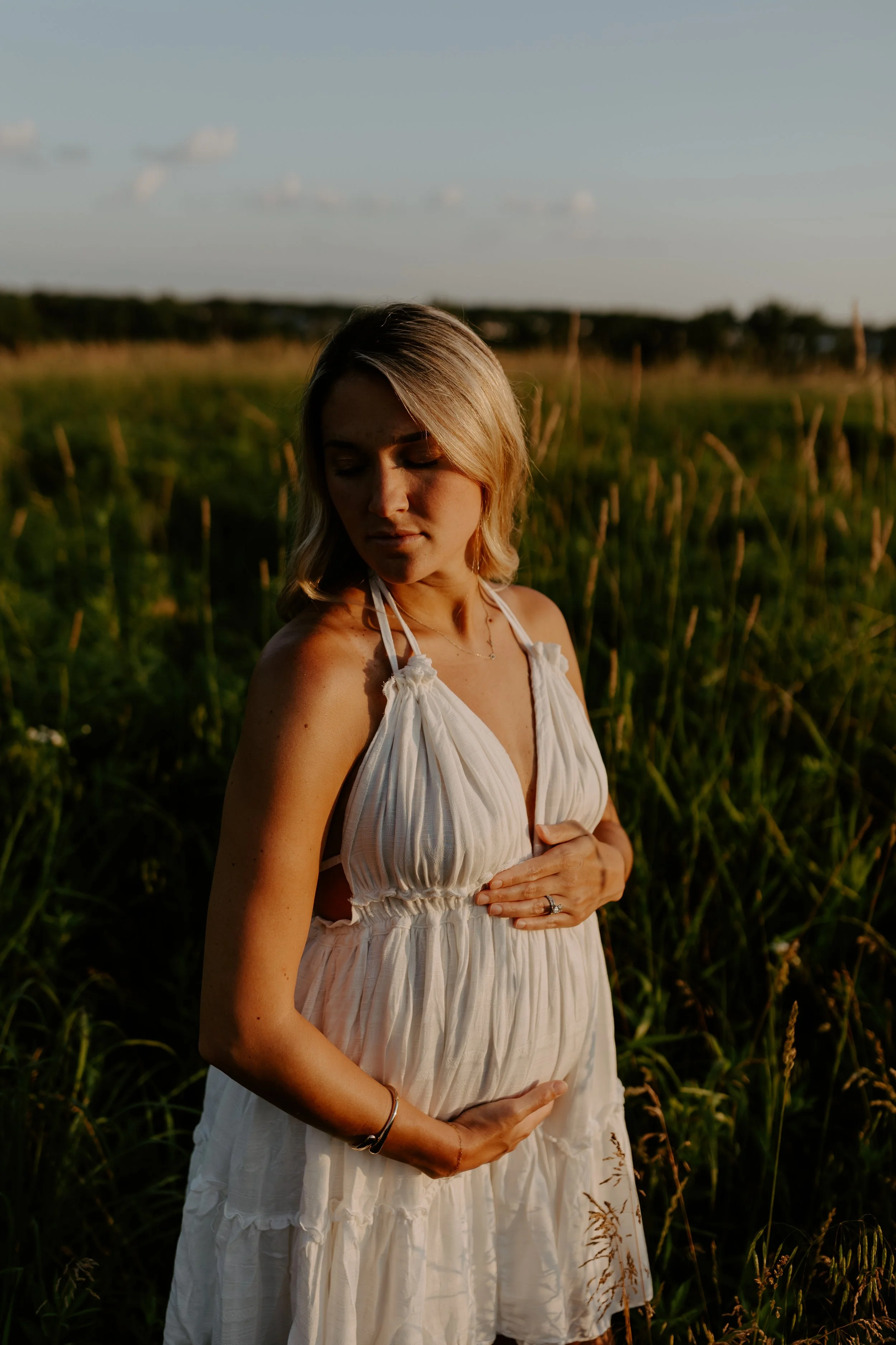 A young woman with blonde hair wearing a white summer dress standing in a grassy field at sunset, with her eyes closed and hands beneath her chest.