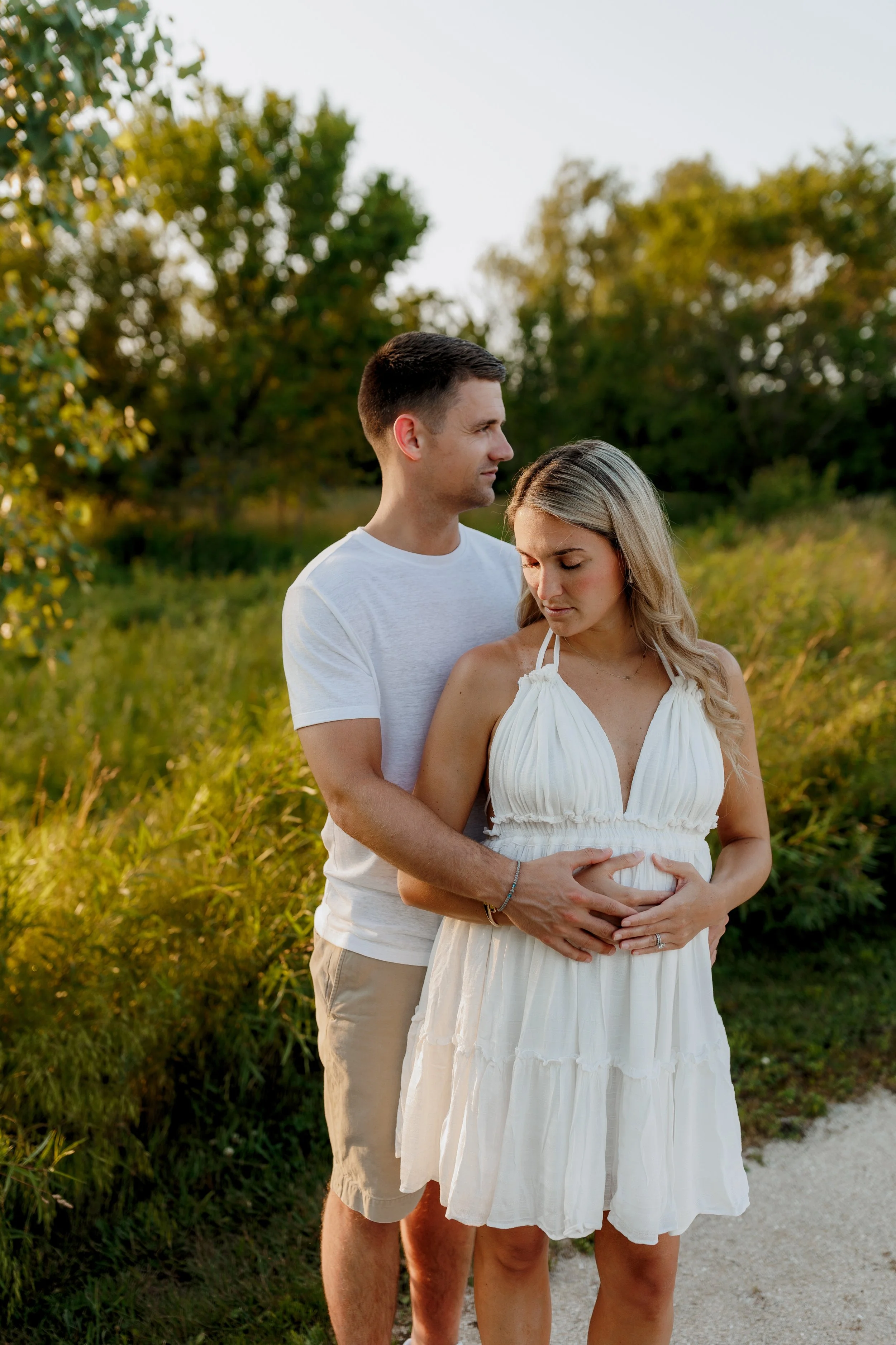 A man and a woman standing outdoors surrounded by greenery. The man is standing behind the woman with his arms around her, holding her belly. They are both wearing light-colored clothing, and the woman appears to be pregnant. The woman is looking dow