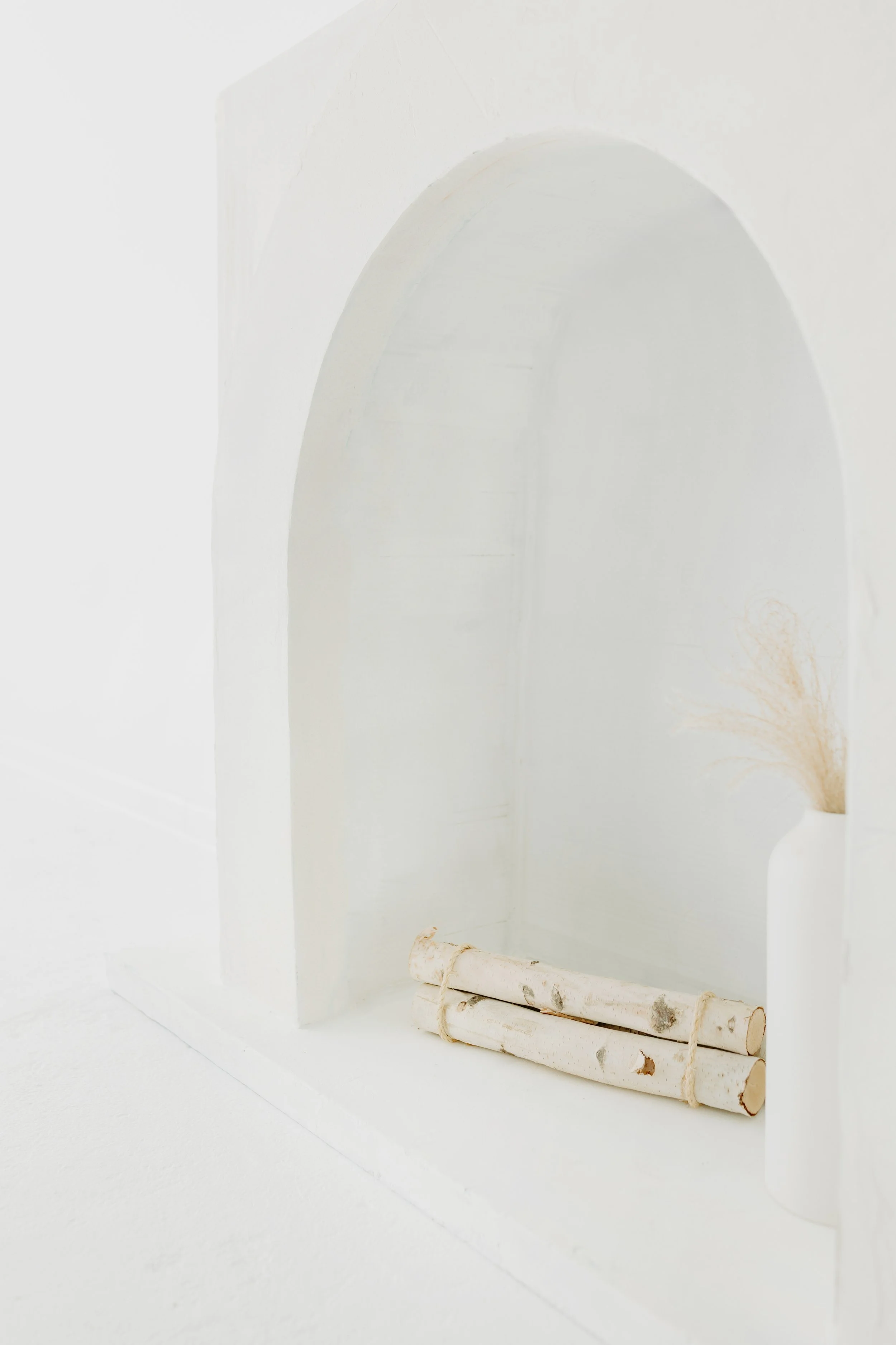 Minimalist white indoor space with a white arch and small white vase containing dried wheat, and two rolled birch logs tied with rope.