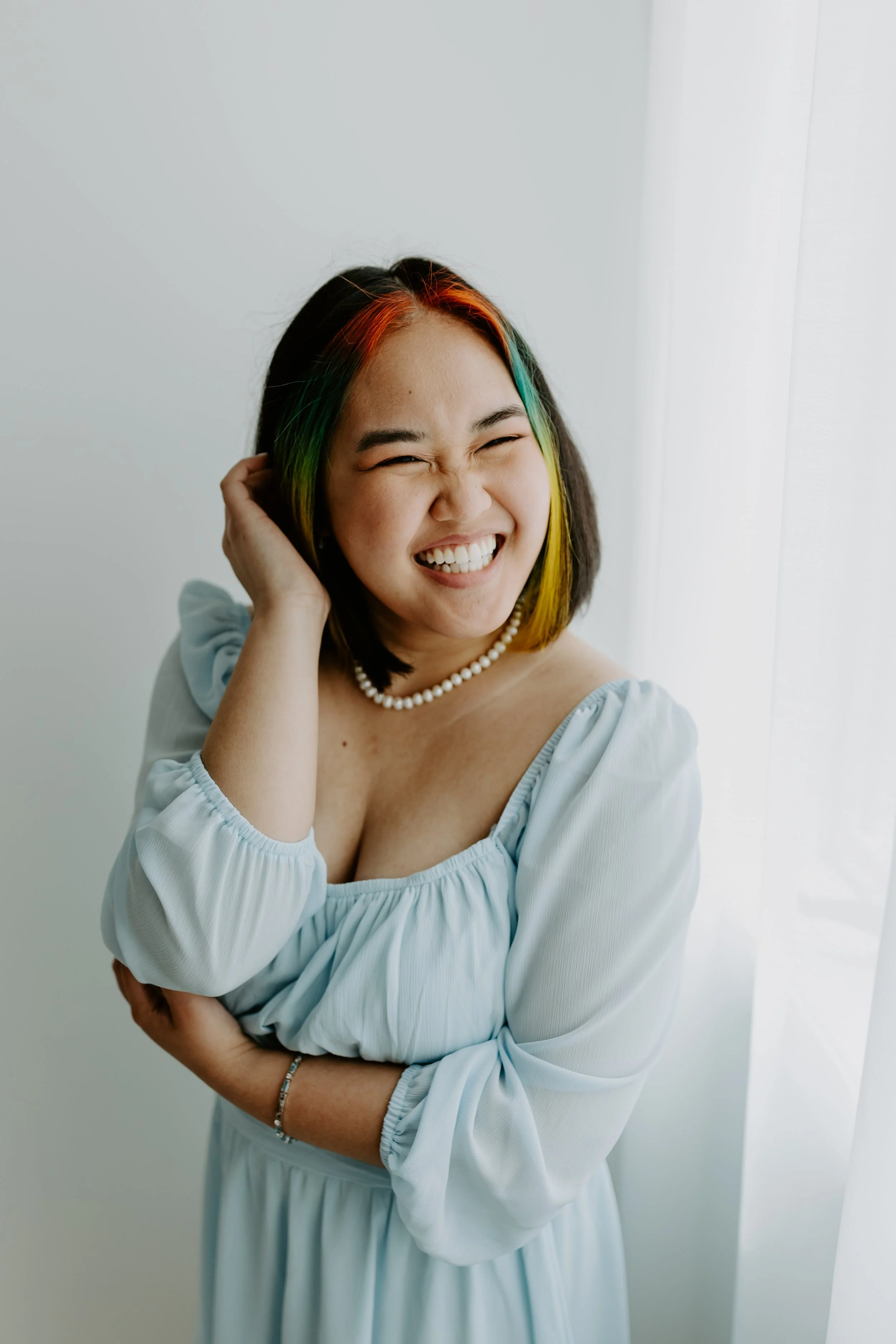 A woman with shoulder-length hair with rainbow colors, smiling and laughing, wearing a light blue dress with puffed sleeves, a pearl necklace, and a bracelet, standing near a white curtain in a bright room.