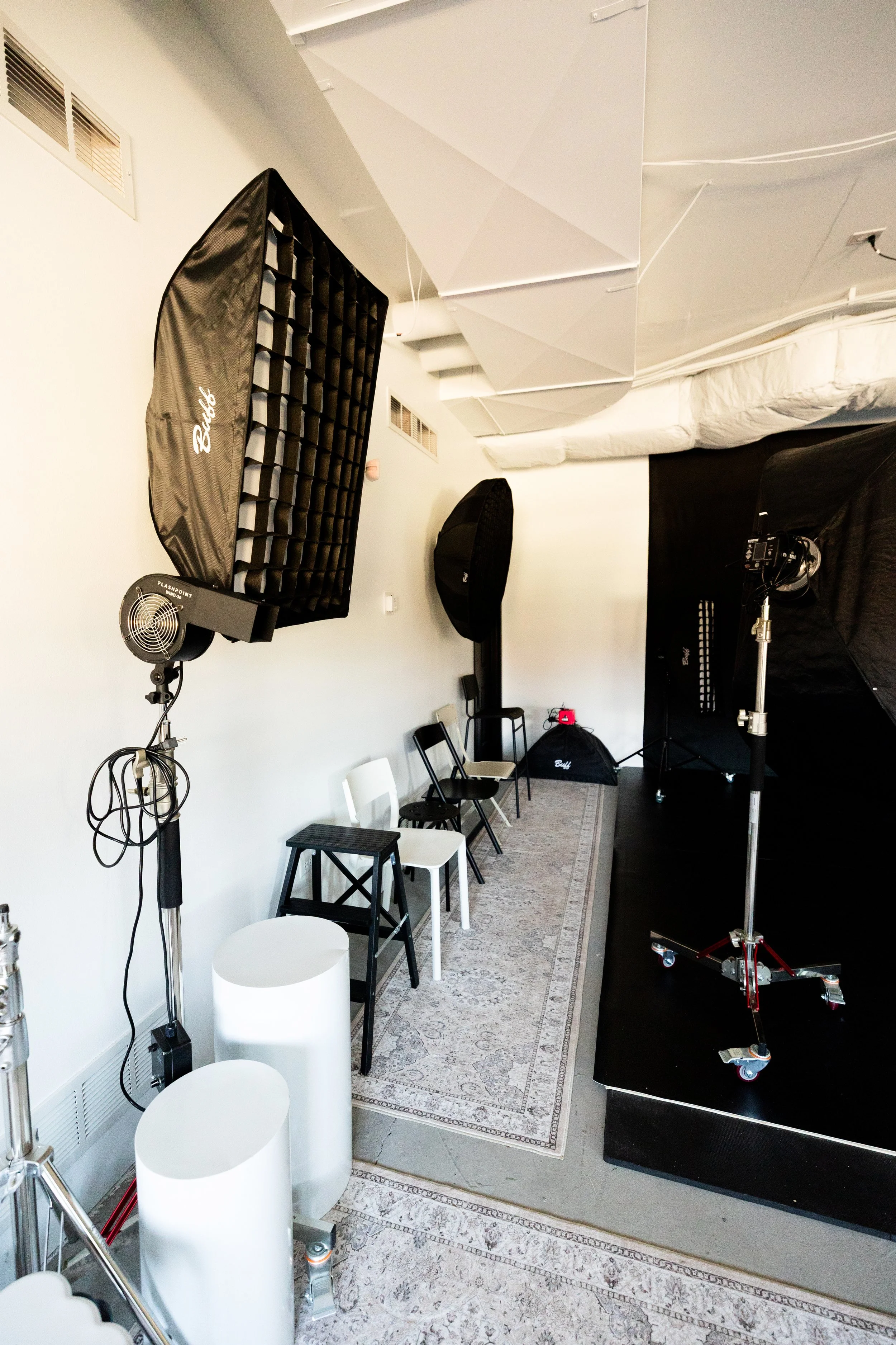 Photography studio with lighting equipment, chairs, a rug, and a raised black platform.