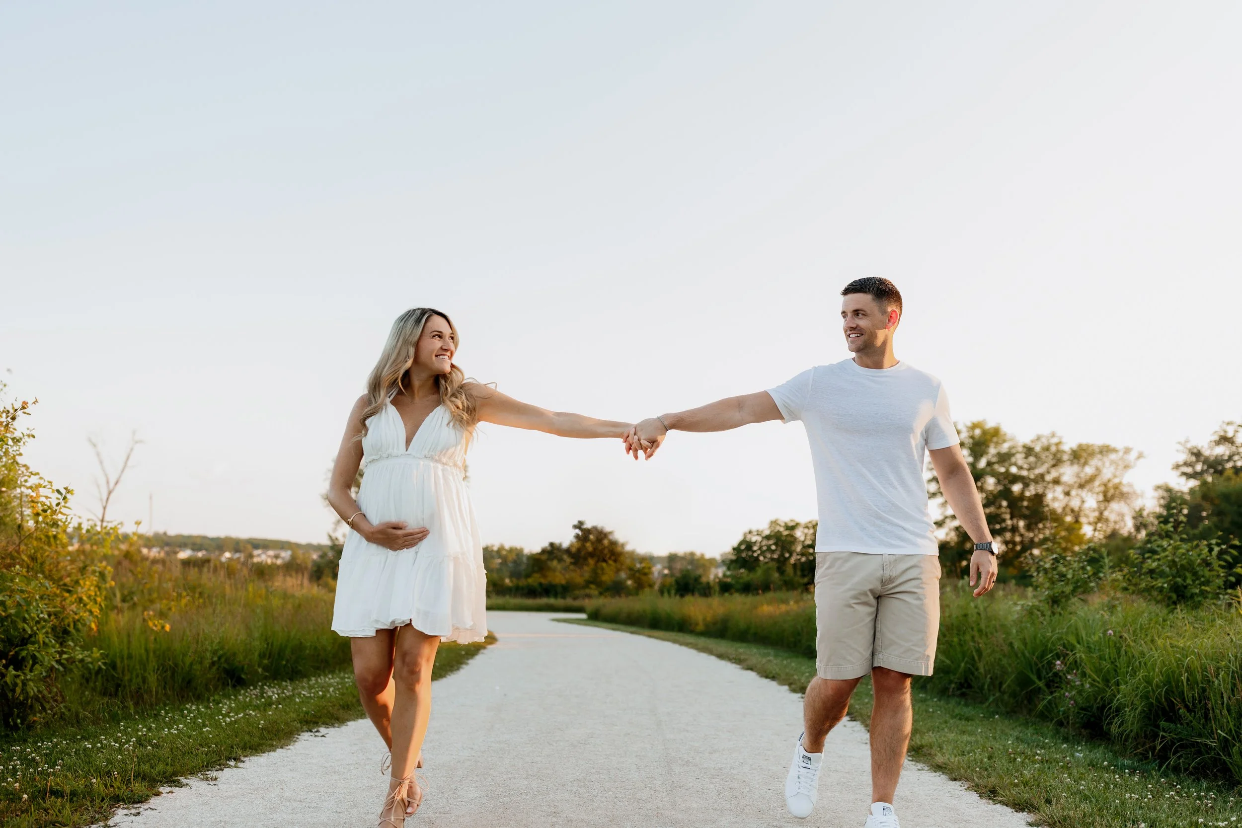 A couple holding hands and smiling while walking along a gravel path in a natural outdoor setting during sunset, with greenery and trees around.