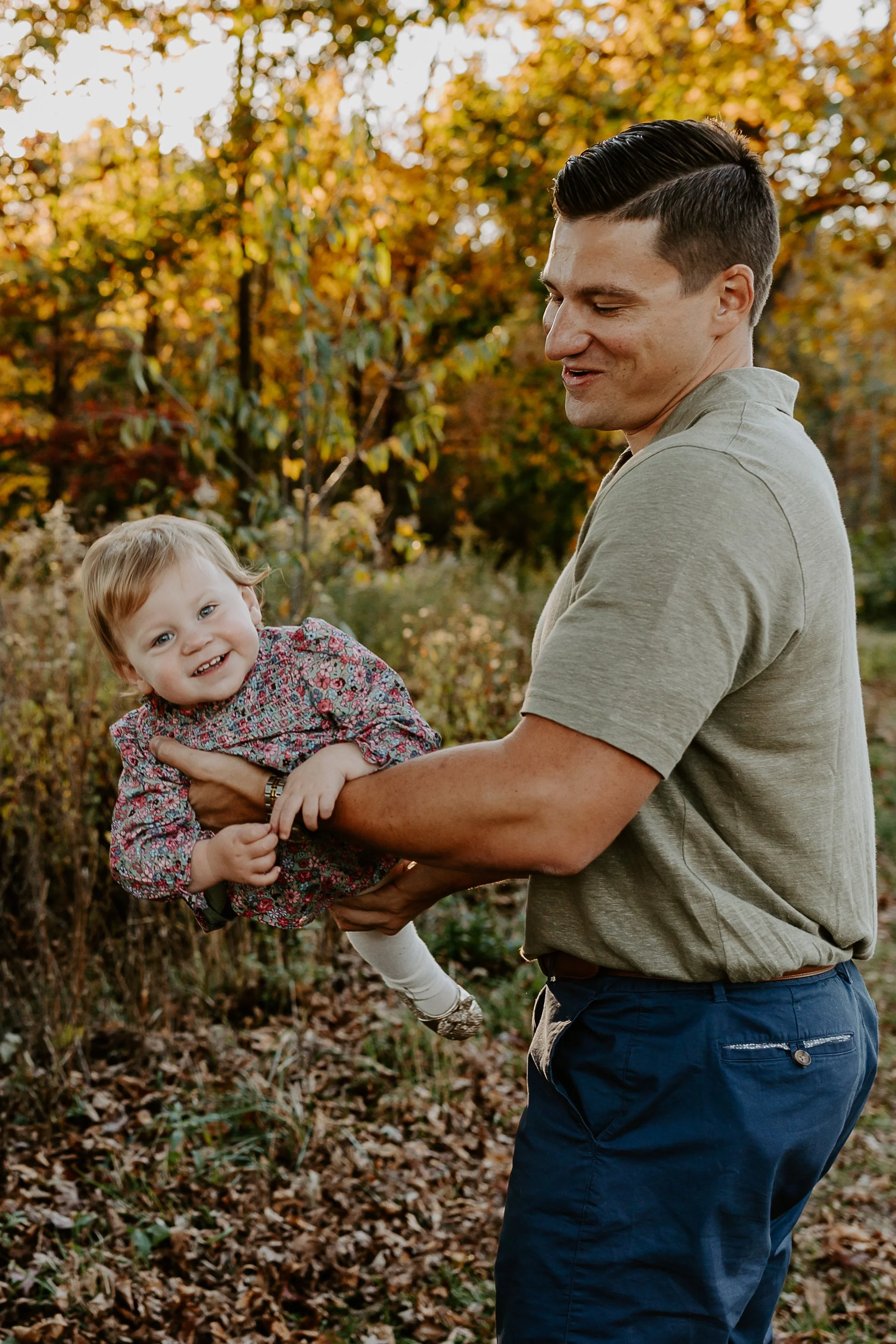 A man in a gray shirt and blue pants holding a smiling young girl with blonde hair in a floral jacket, outdoors during autumn with trees and fallen leaves.