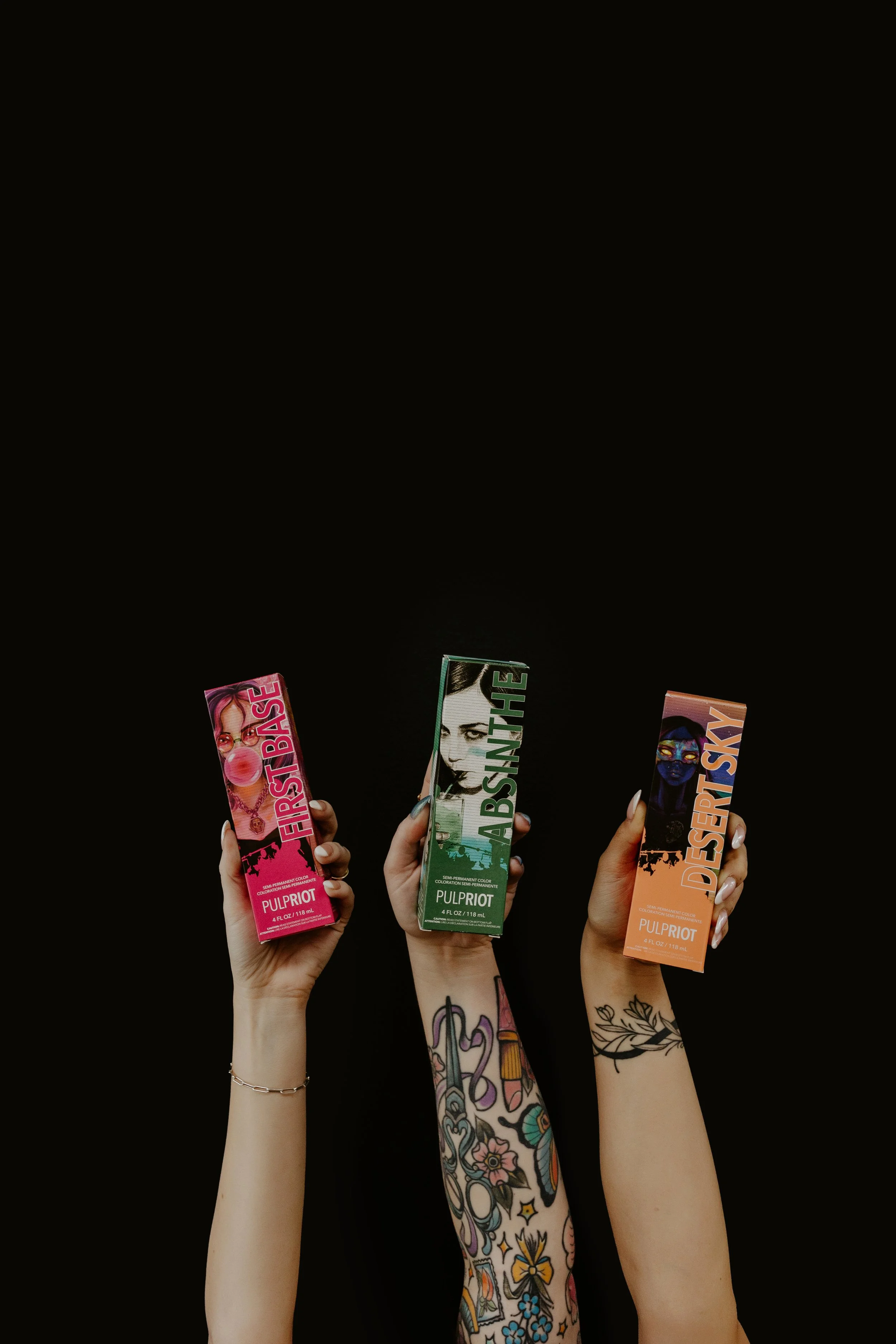 Three hands holding hair dye boxes against a black background. The left hand has a pink box labeled "First Base" with a woman blowing bubble gum. The middle hand has a green box labeled "Absinthe" with a woman's face. The right hand has an orange box