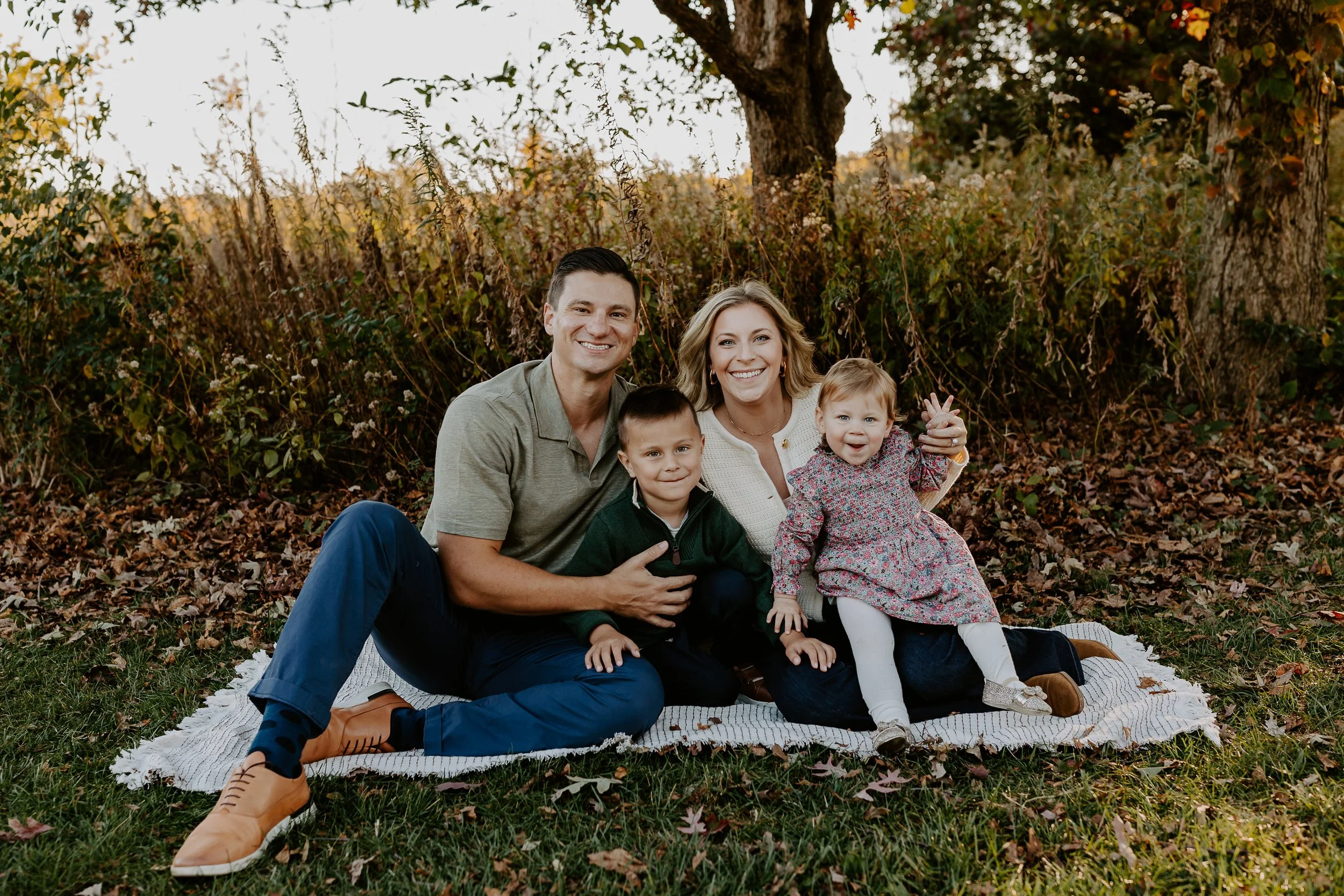 A family of four sitting on a blanket outdoors among autumn leaves, smiling at the camera. The background features trees and fall foliage.