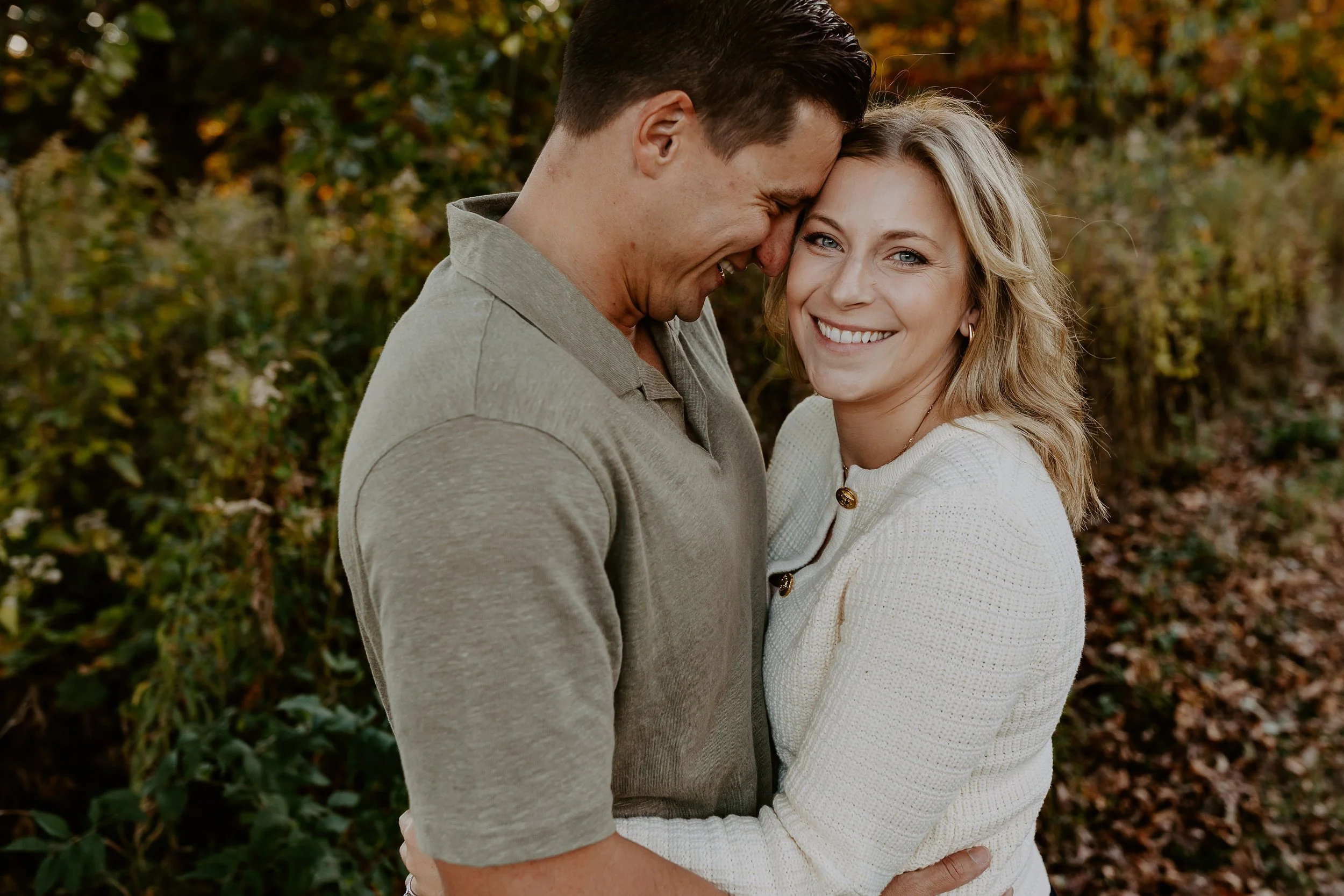 A couple hugging outdoors in fall, smiling with their foreheads touching among trees with autumn leaves.