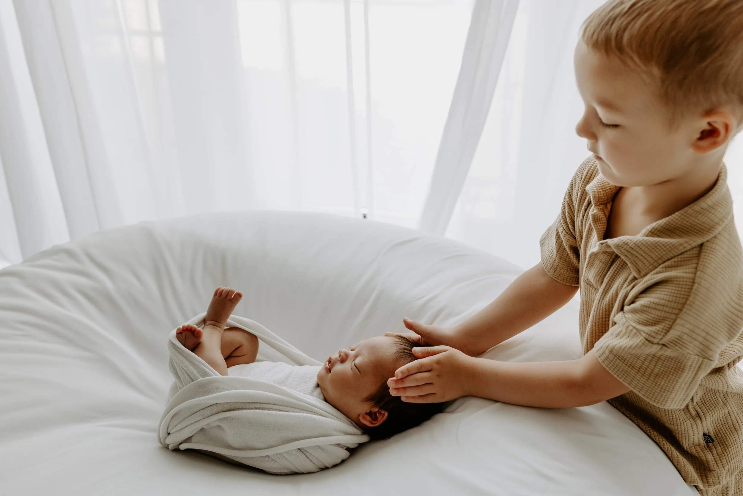 A young boy gently holding and playing with two small babies on a bed in a bright room with white curtains.