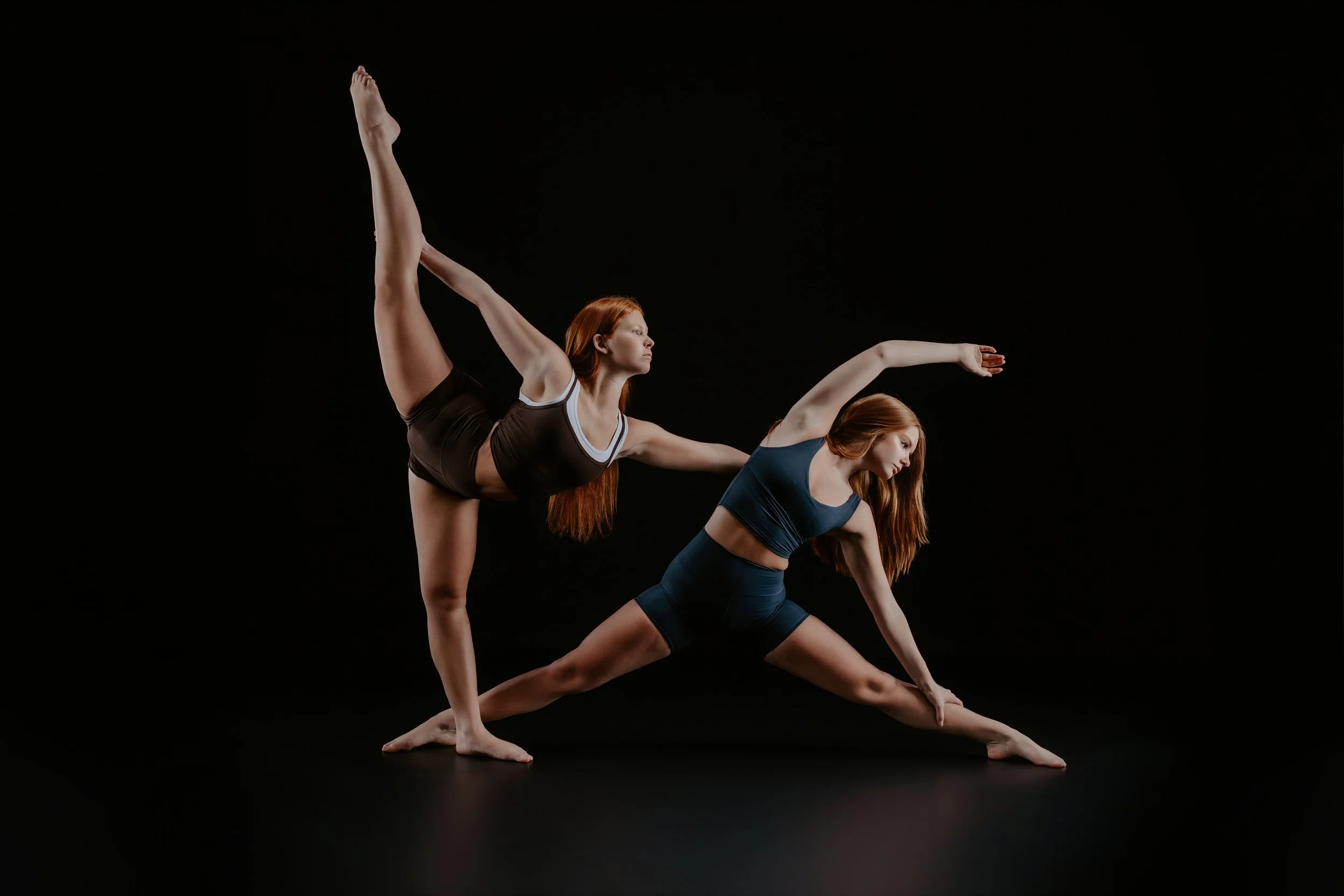 Two women practicing yoga together, performing balancing poses on a black background.