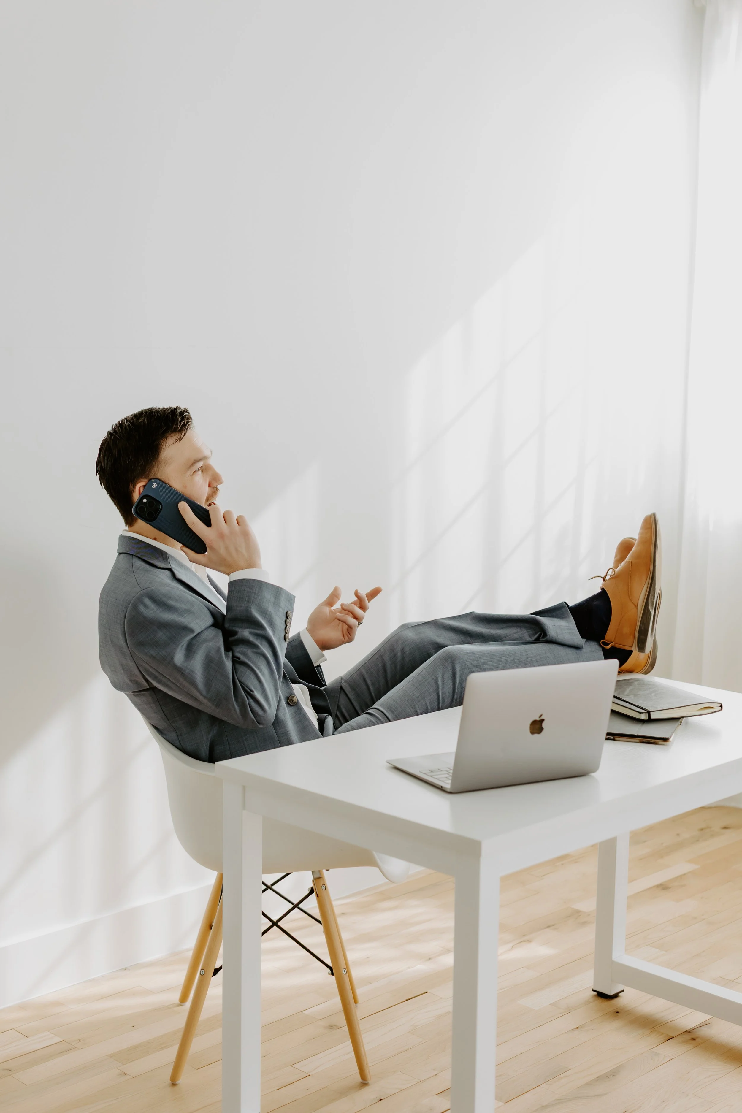 Businessman in a gray suit sitting at a desk, talking on a phone, with his feet up on the desk.