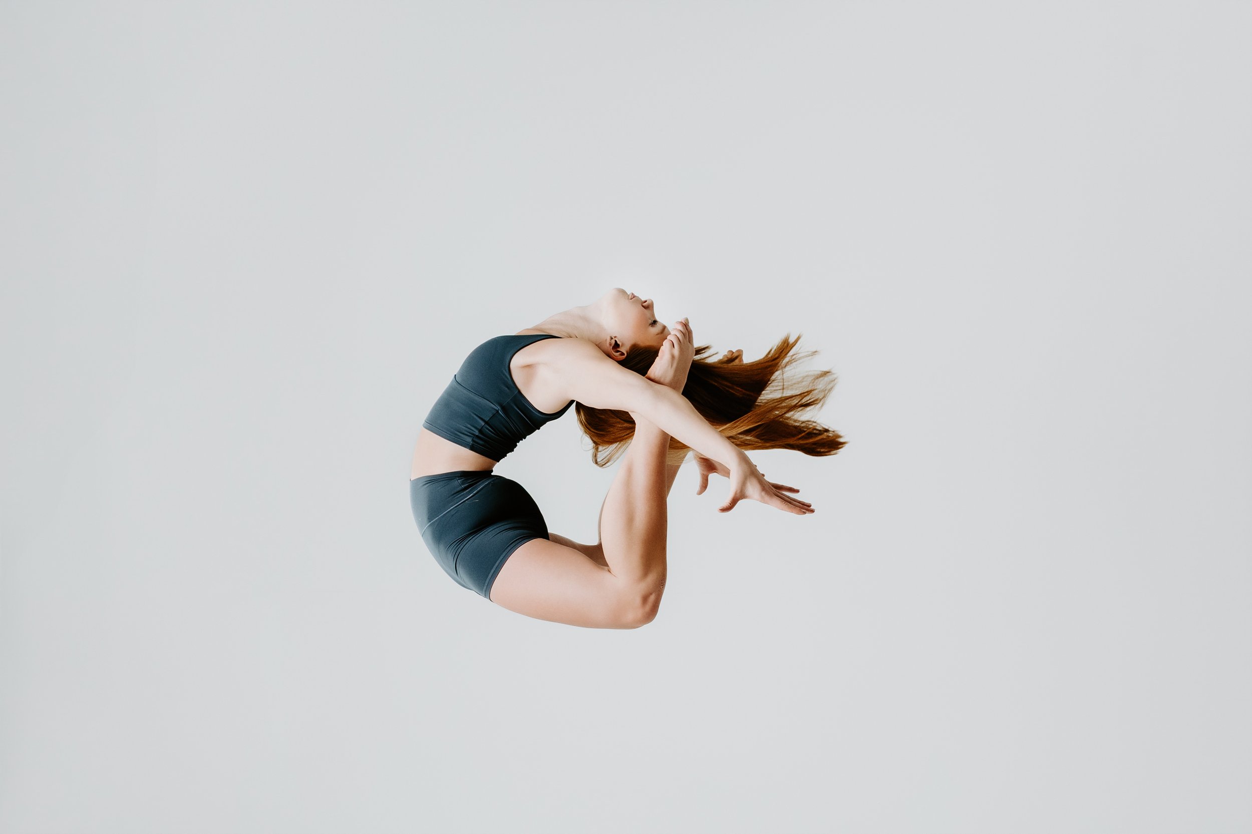 A woman performing a yoga pose on a plain light gray background, wearing a black sports bra and shorts.