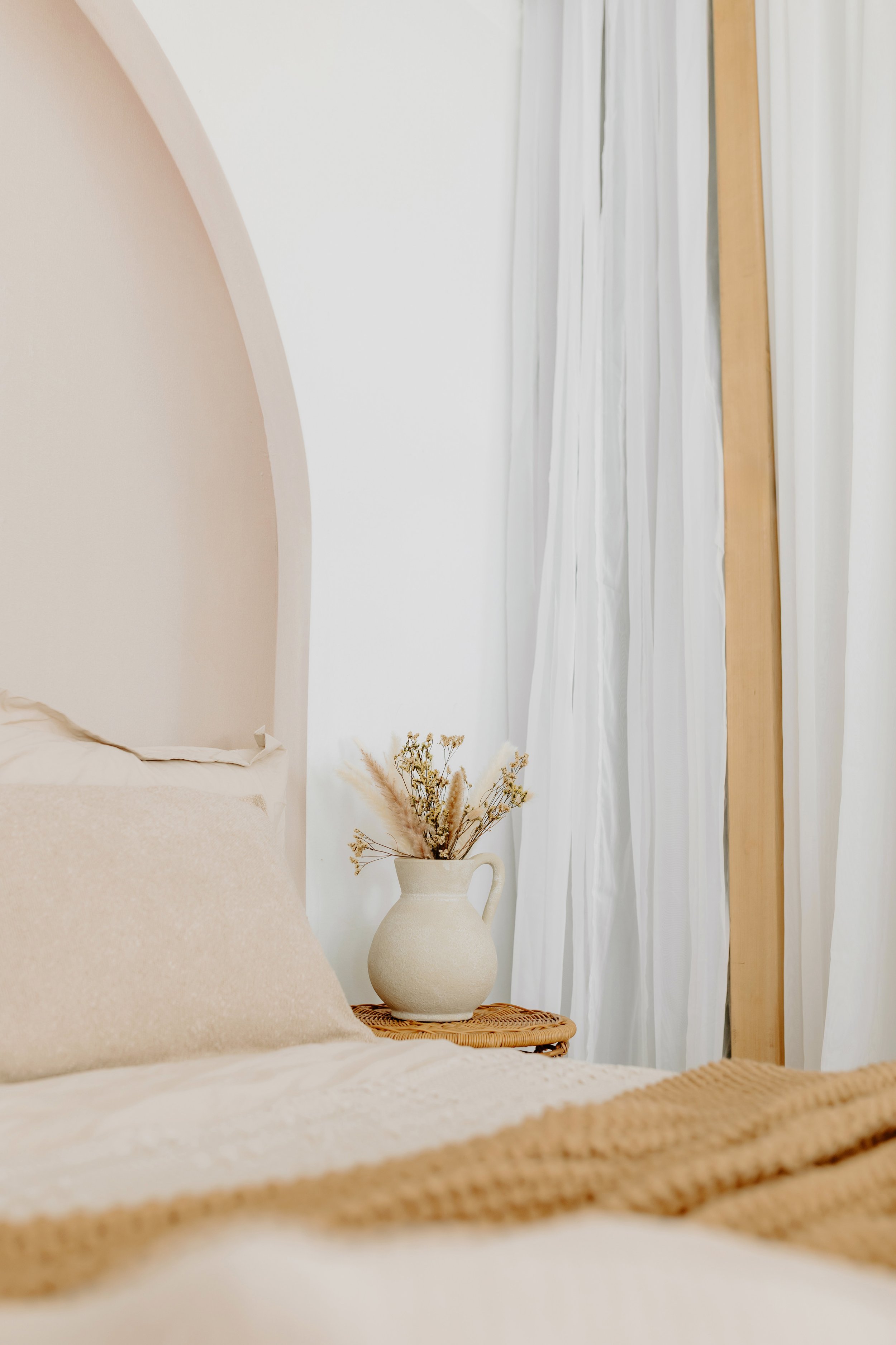 A cozy bedroom with a beige headboard, neutral-colored bedding, and a nightstand with a white vase filled with dried flowers, with white curtains in the background.
