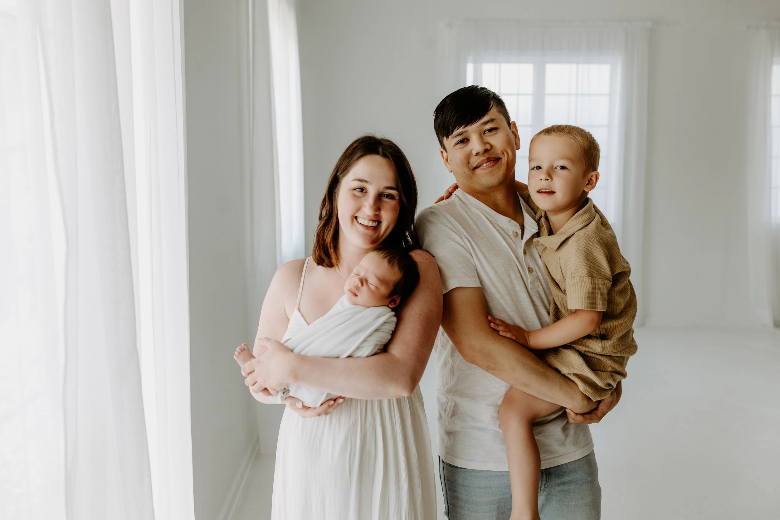 A family of four, including a mother, father, and two young children, standing together in a bright room with white curtains and windows.