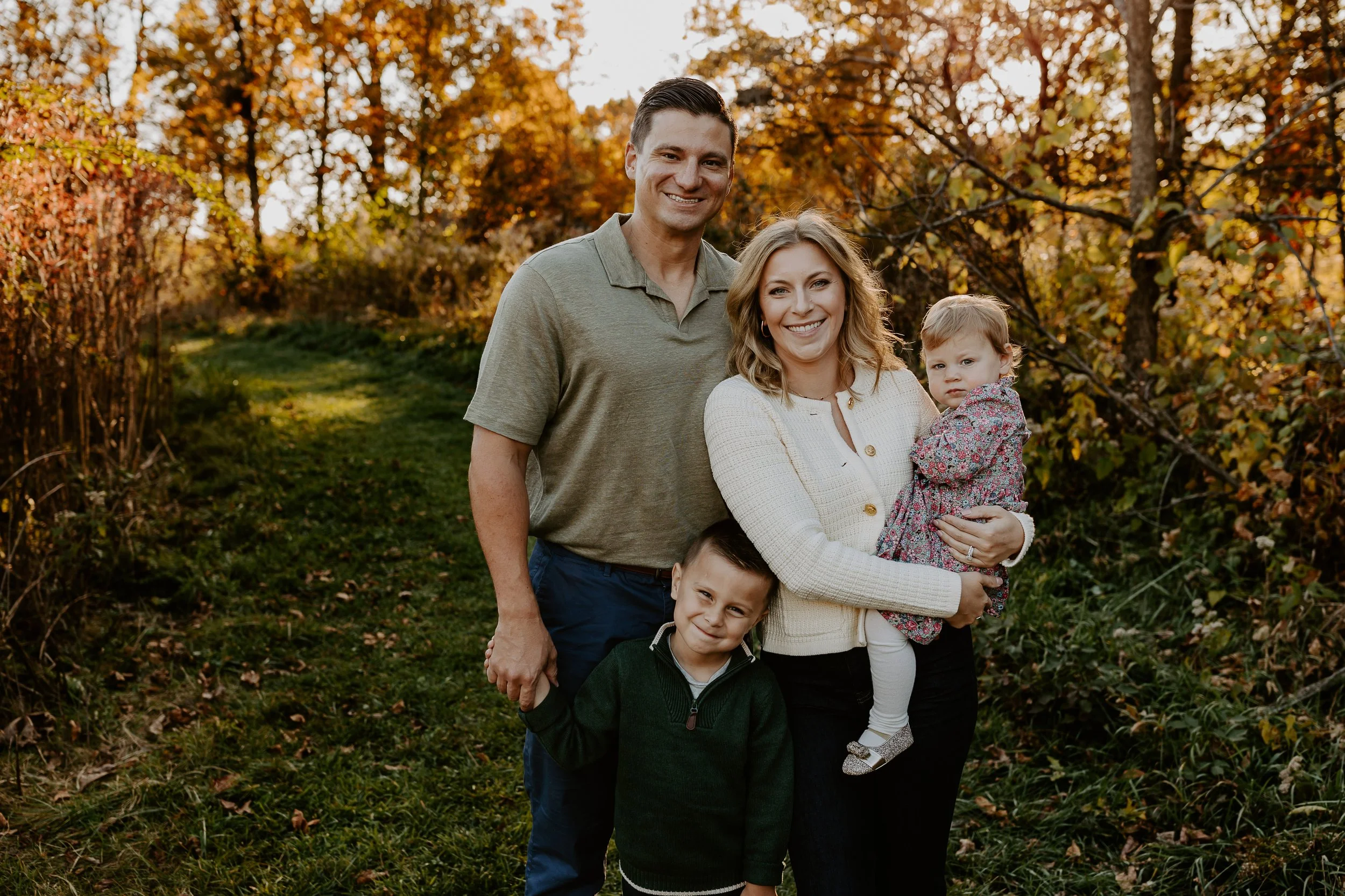 A family of five people smiling and posing outdoors during fall, with colorful autumn trees in the background.