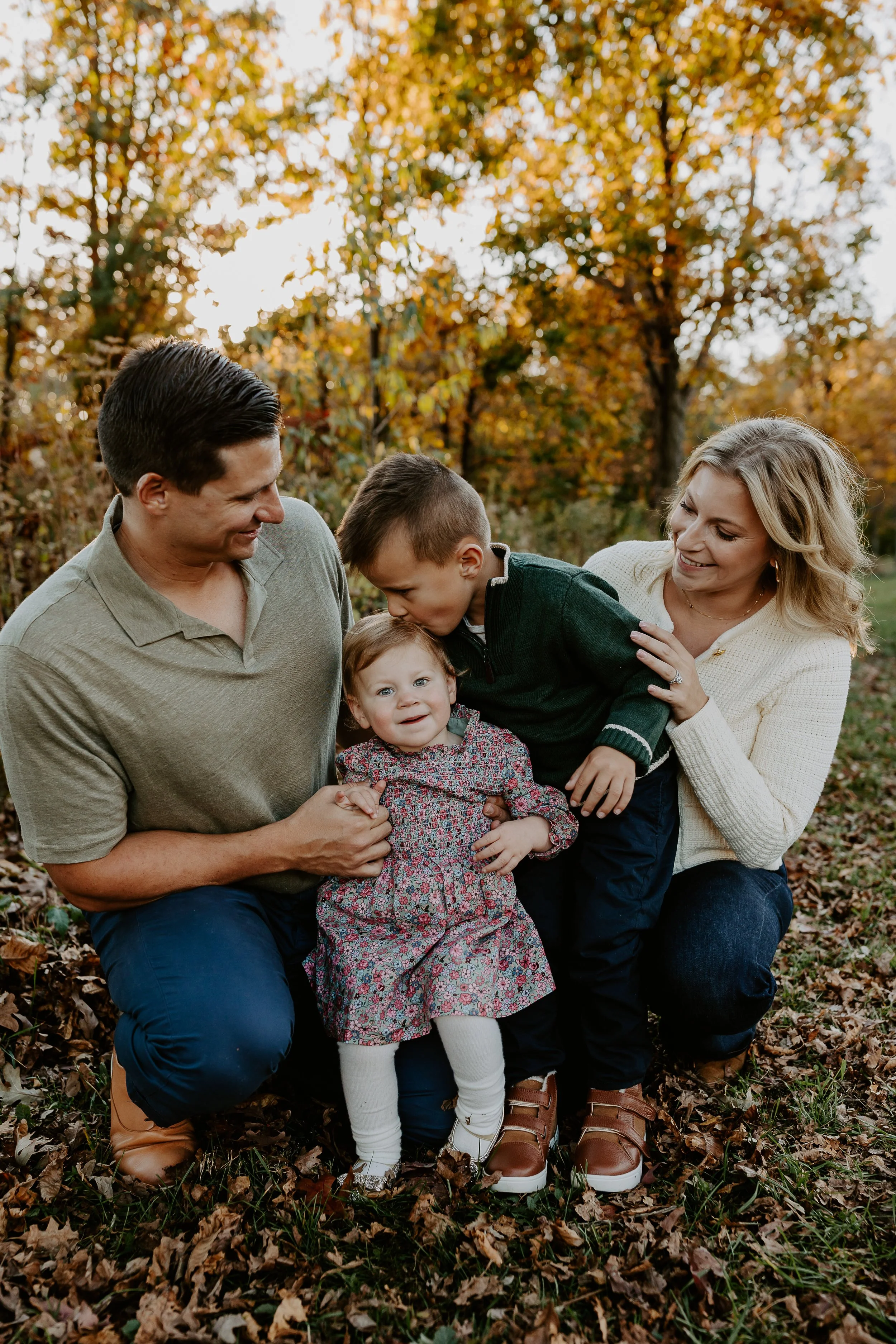 A family of five, including two adults and three children, is outdoors during fall, surrounded by autumn leaves and trees with orange and yellow foliage, smiling and enjoying time together.