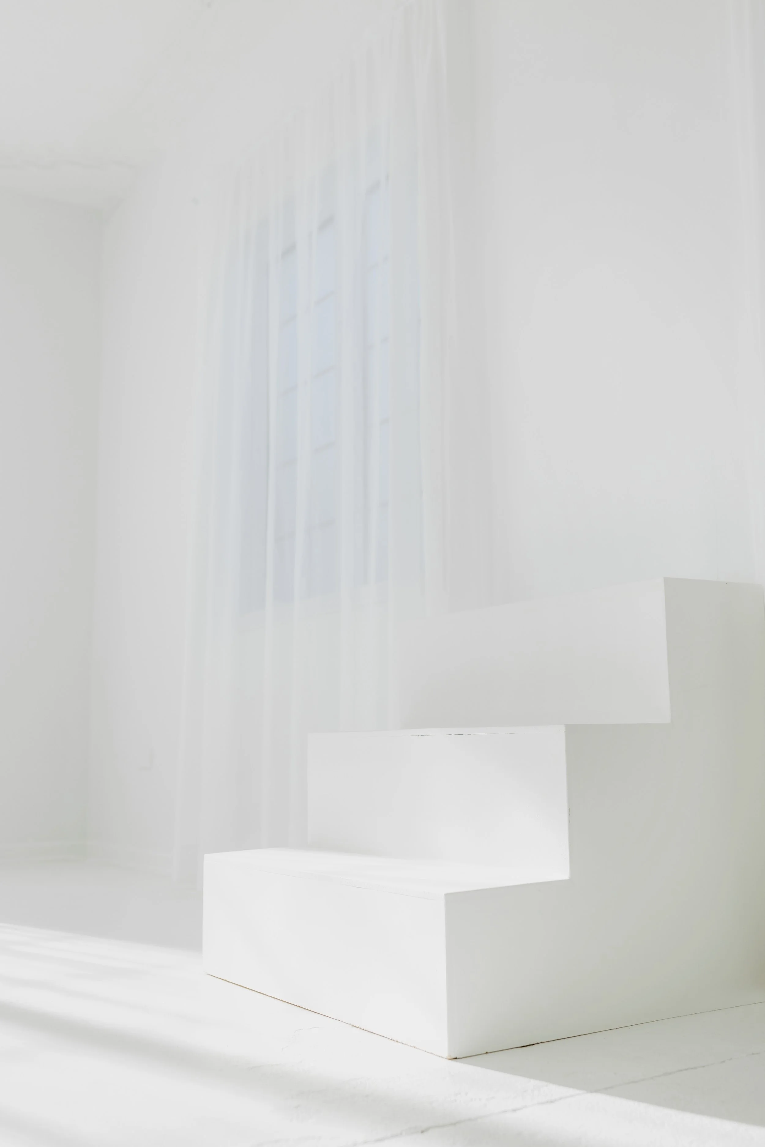 Minimalist white staircase near a window with sheer white curtains in a bright, airy room.