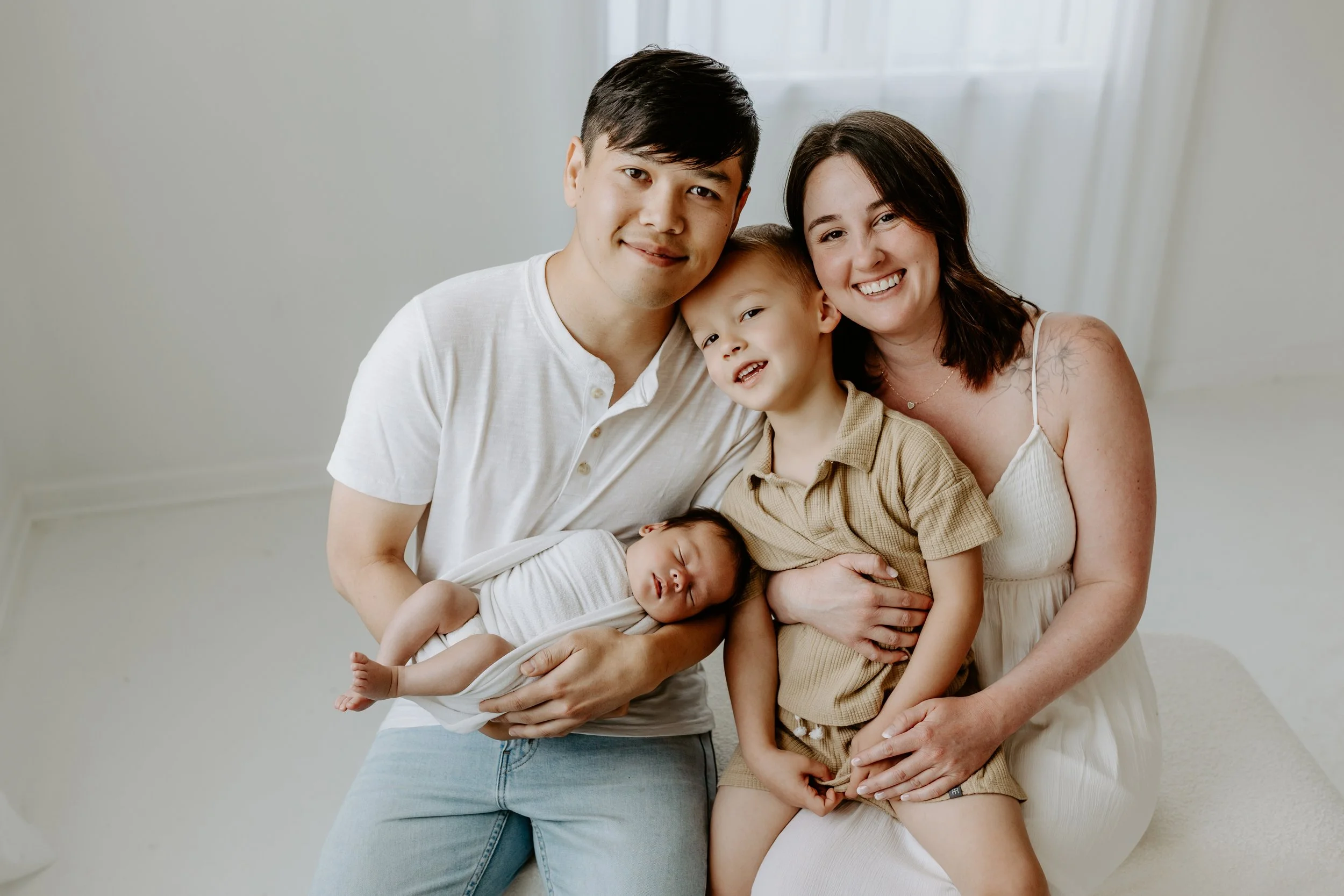 A happy family of five, including a man, woman, two young children, and a newborn, posing indoors with smiles.