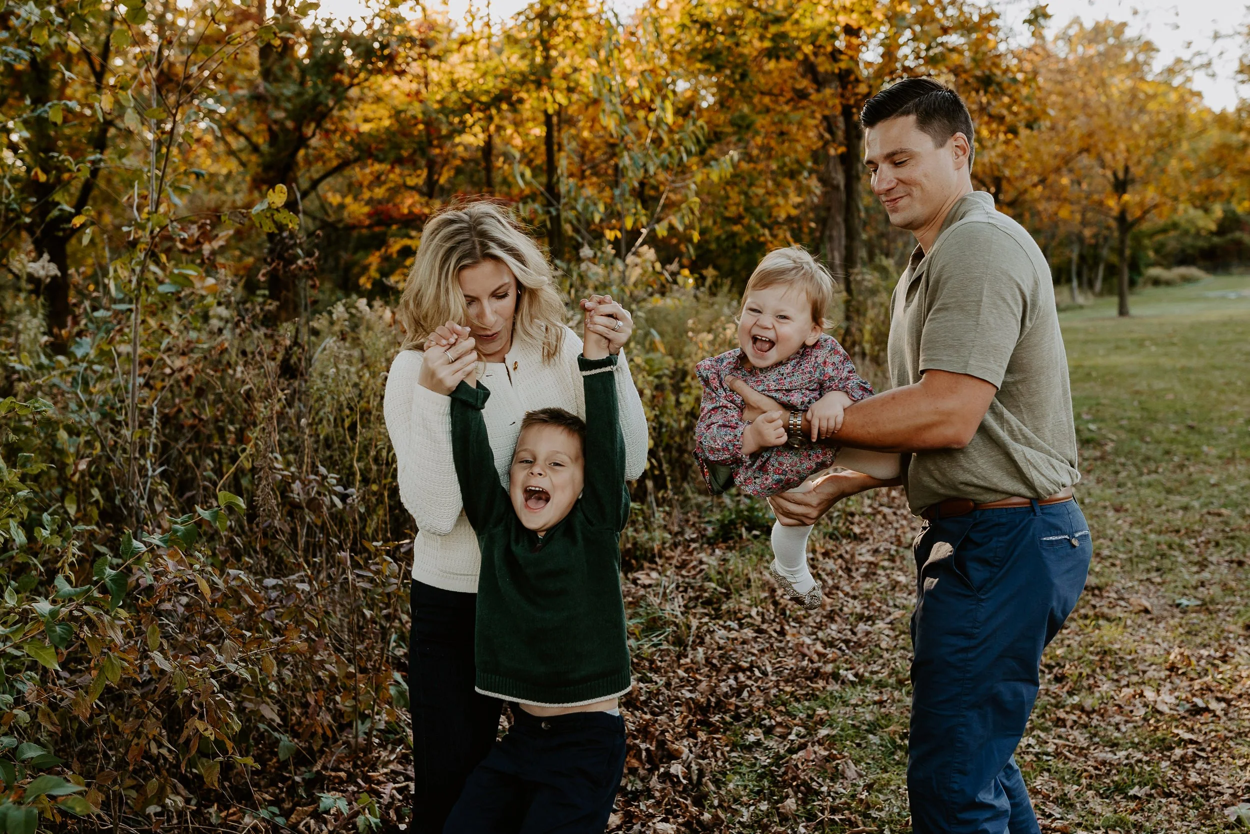 A family of four, including two young children, is enjoying a walk outdoors in autumn with colorful fall foliage in the background. The mother, father, and children are smiling and having fun.