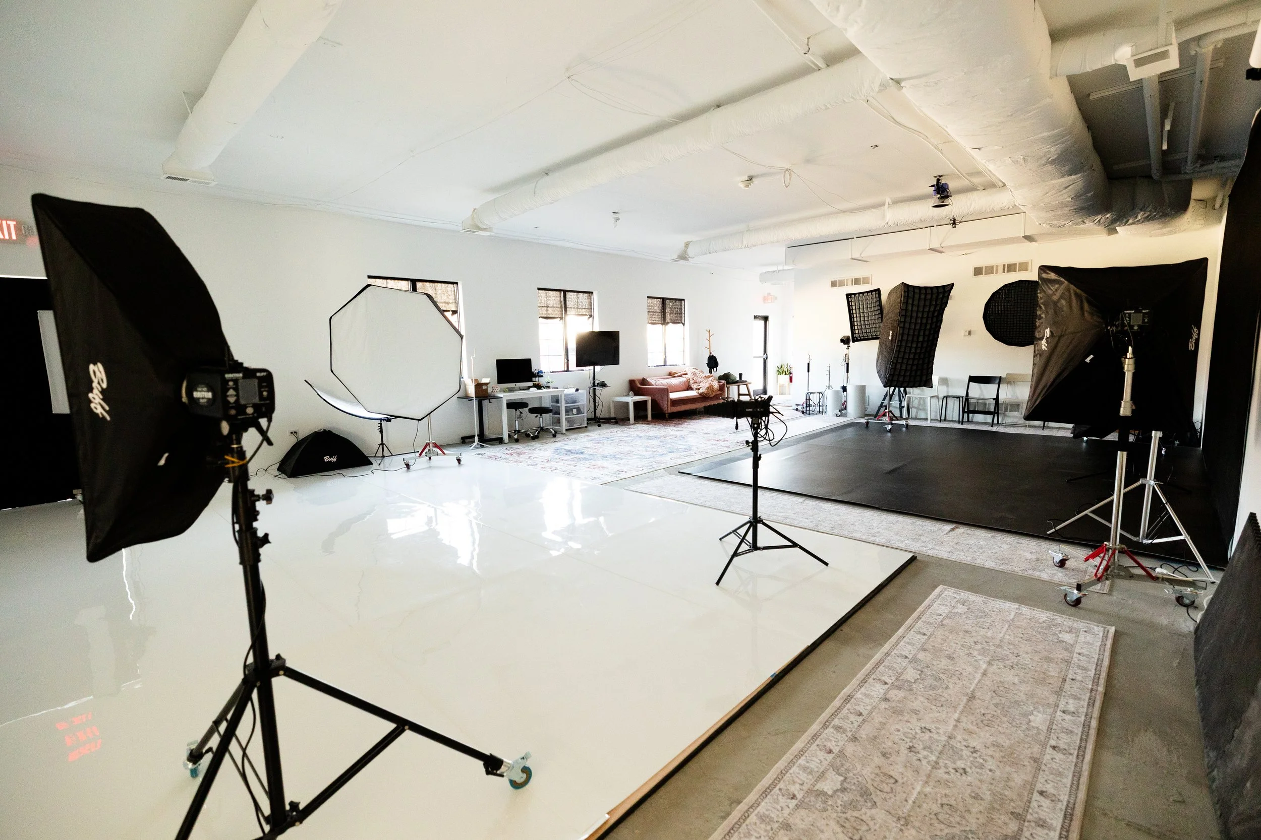 Empty professional photography or video studio with lighting equipment, umbrellas, black backdrop, a sofa, and desks, in a spacious room with white walls and large windows.