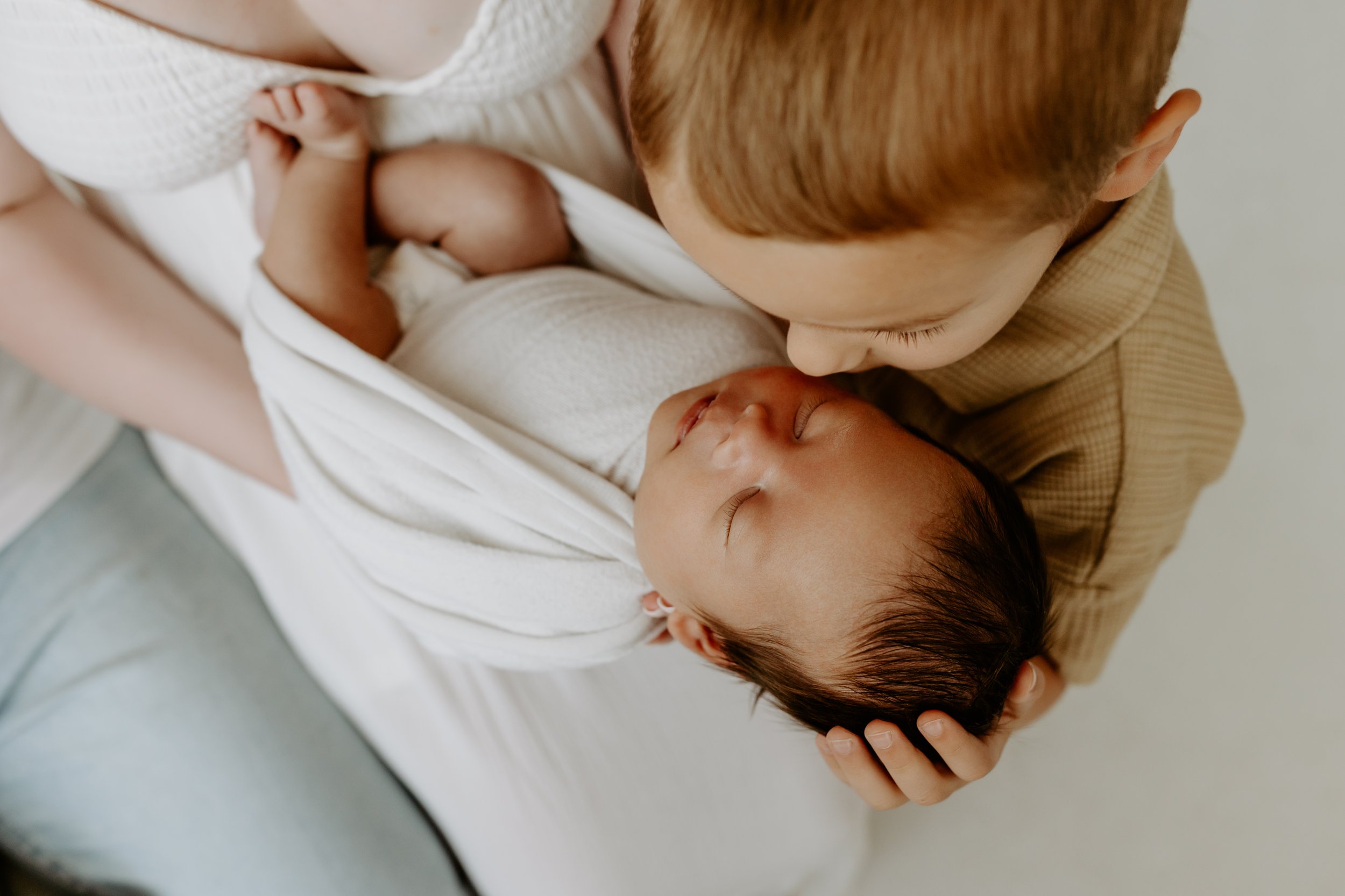 A person with light skin and reddish-brown hair is gently kissing a newborn baby who is wrapped in a white blanket, while another person with darker skin and dark hair holds the baby's head.