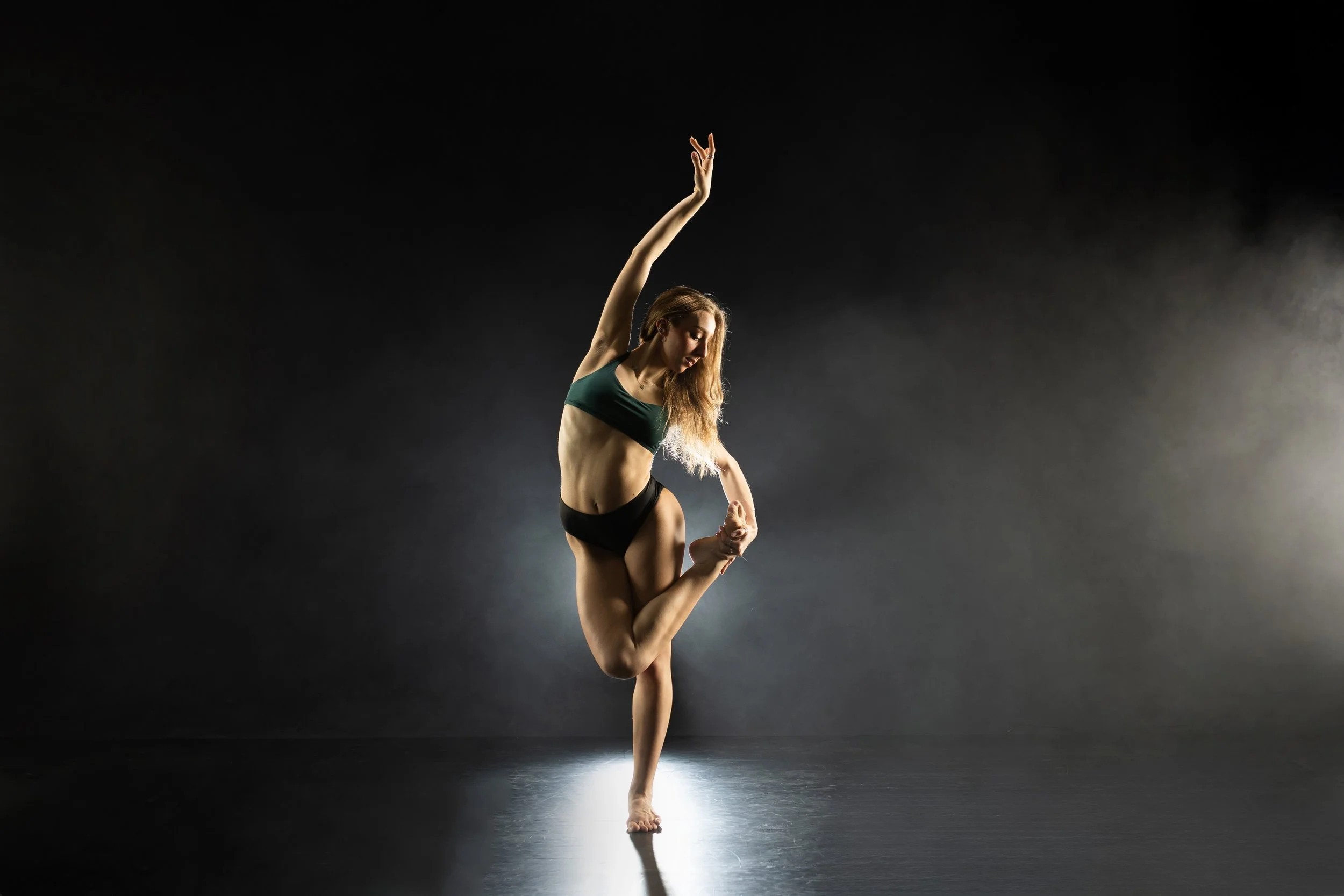 A woman with long blonde hair dancing alone in a dark studio, performing a pose where she balances on one leg with her other leg bent and held by her hand, one arm extended upward.
