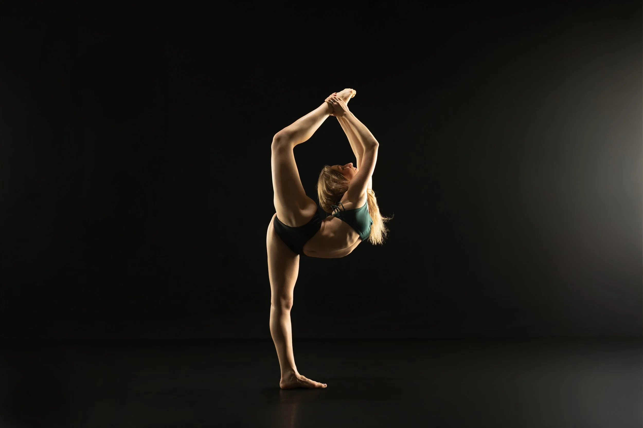 A woman on a black background performing a standing backbend yoga pose with one leg lifted and held by her hands.