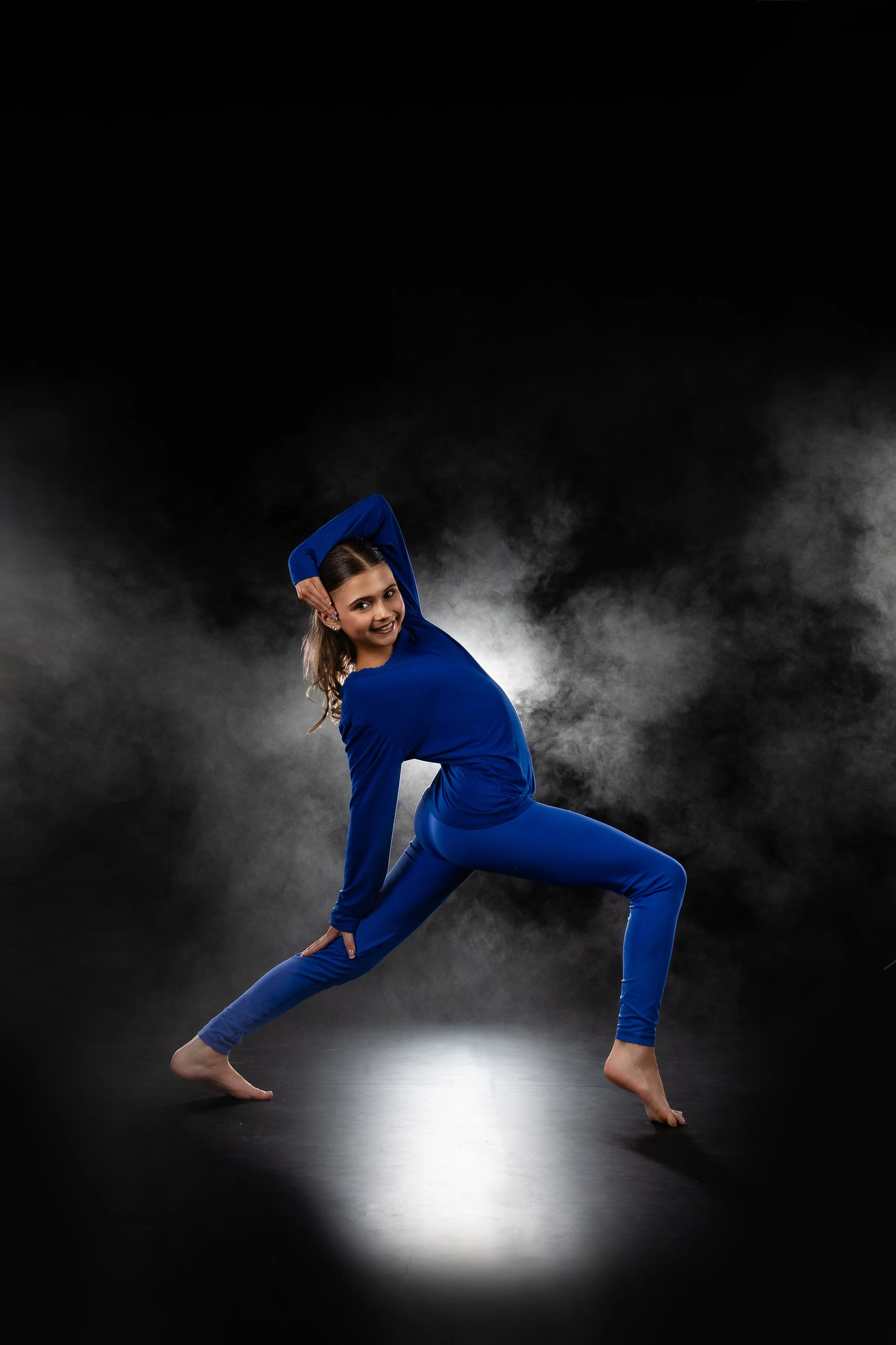 A woman in a blue outfit poses in a dance or yoga studio with smoky background lighting.