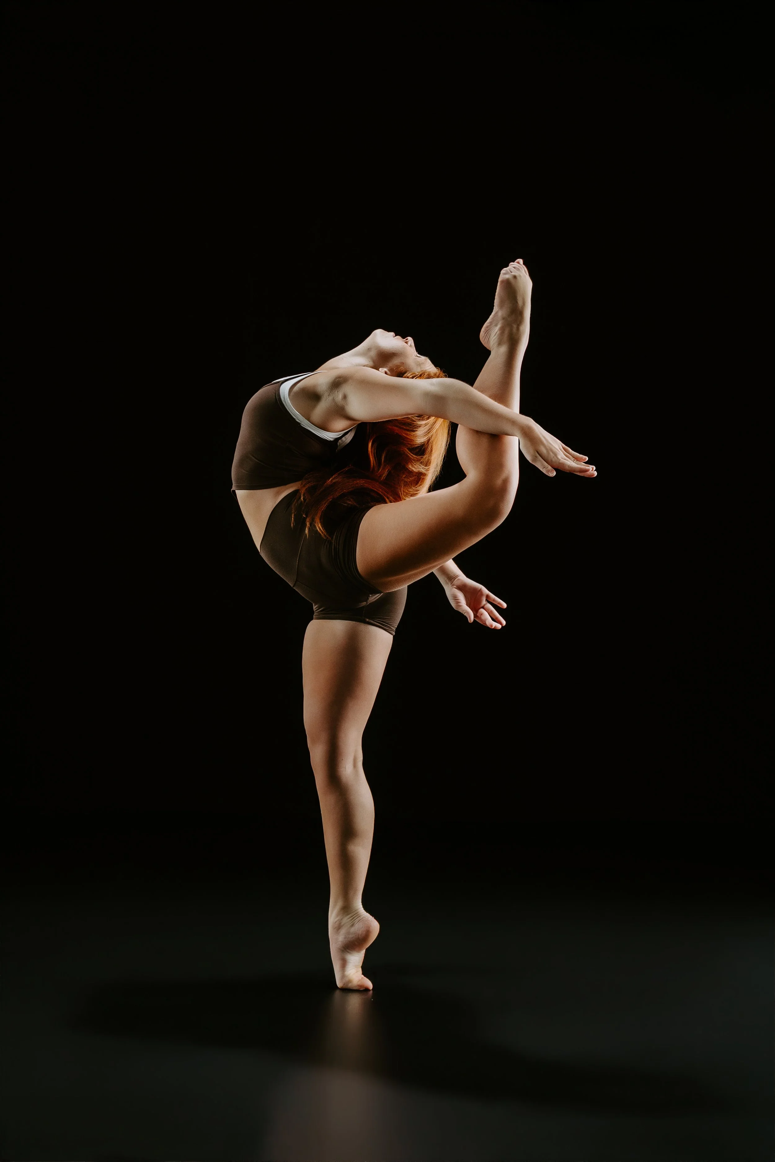 A dancer with red hair performing a balancing pose on a black background.
