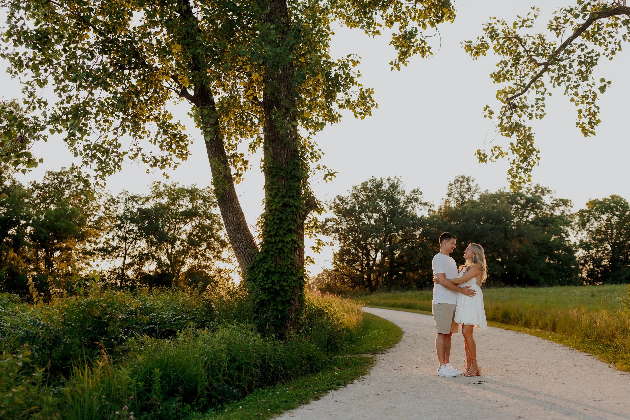 A couple standing together on a park path during sunset, holding each other and smiling. There are tall trees with green leaves and a grassy area surrounding them.