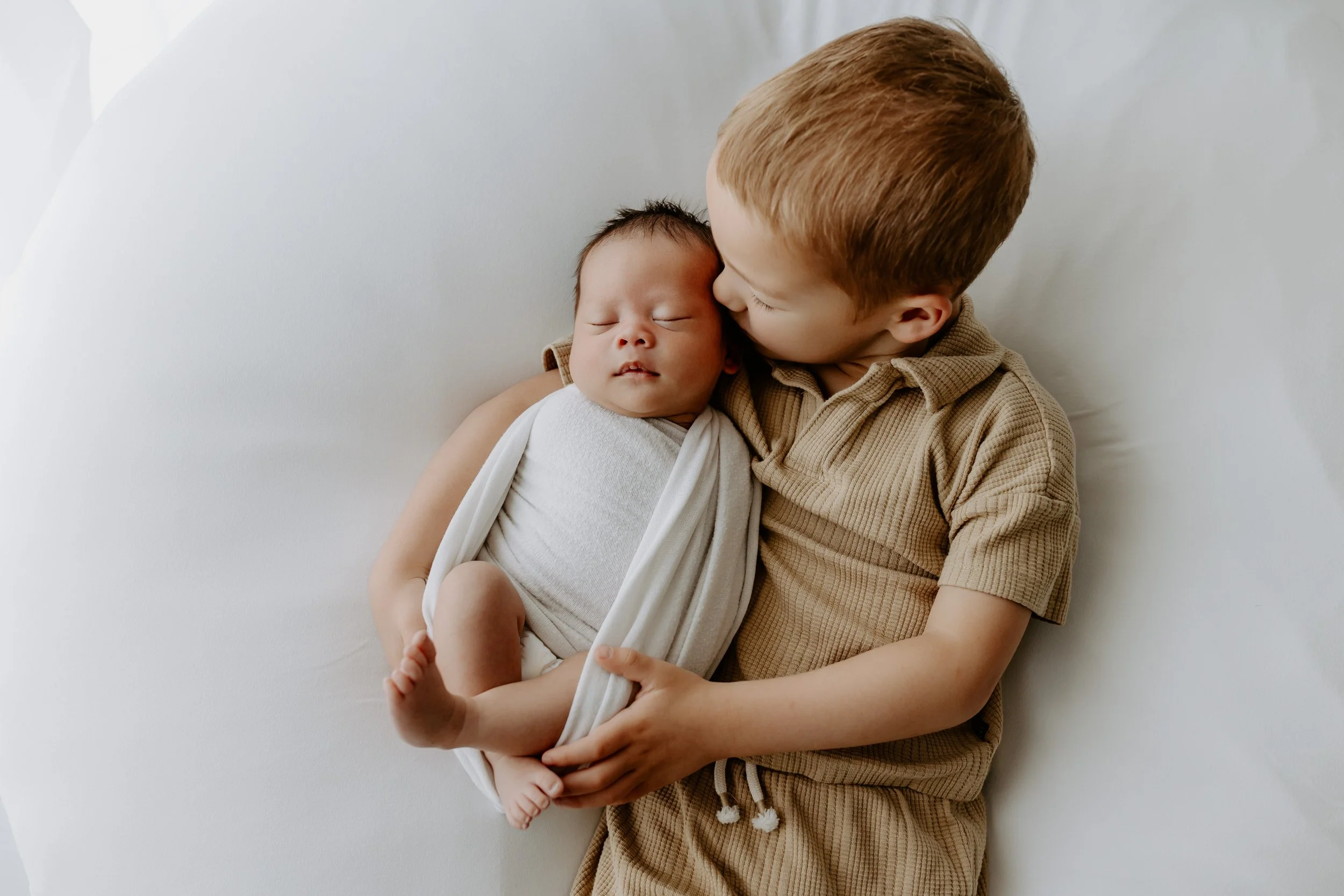 A young boy with red hair holding and kissing a sleeping newborn baby wrapped in a white blanket.