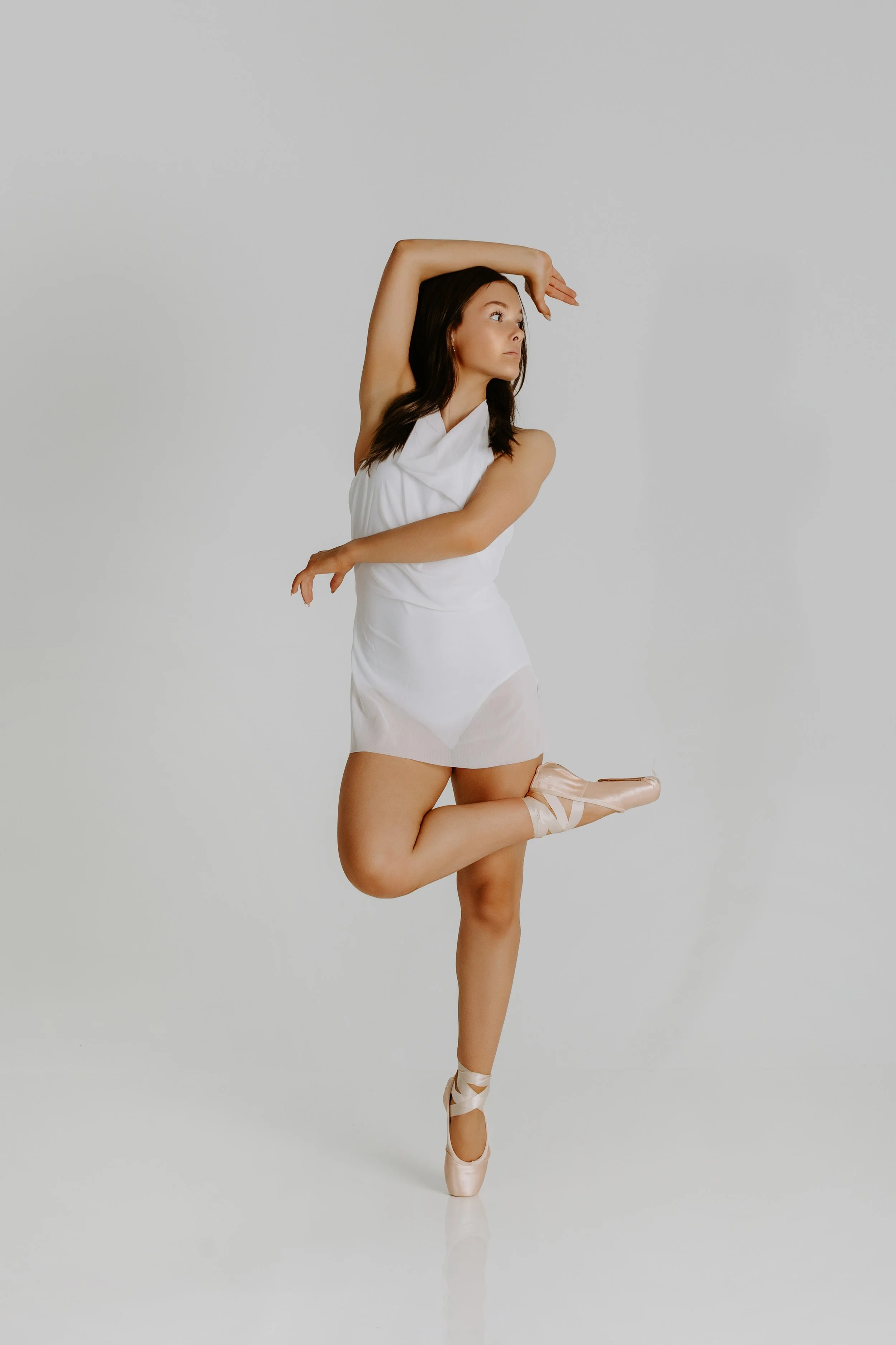 A young woman in a white dress and ballet shoes is standing on one leg, with her other leg bent at the knee and raised behind her. She is balancing on her toes, dancing, with one arm bent and resting on her head and the other arm extended downward. S