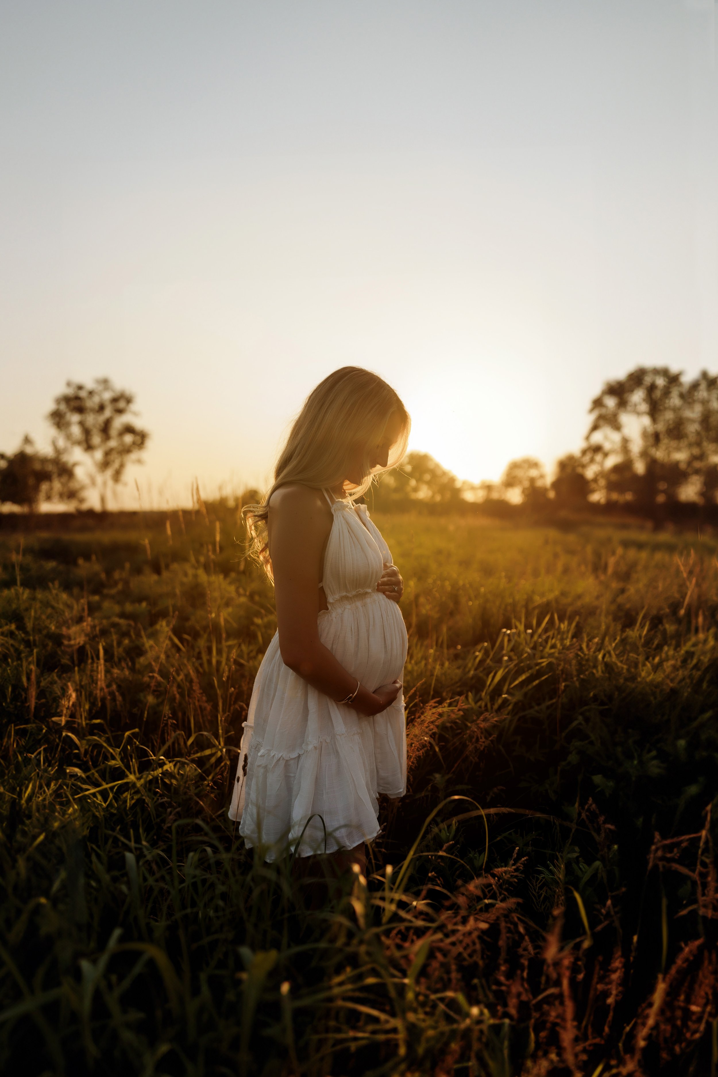 A pregnant woman in a white dress stands in a field at sunset, holding her belly and looking down.
