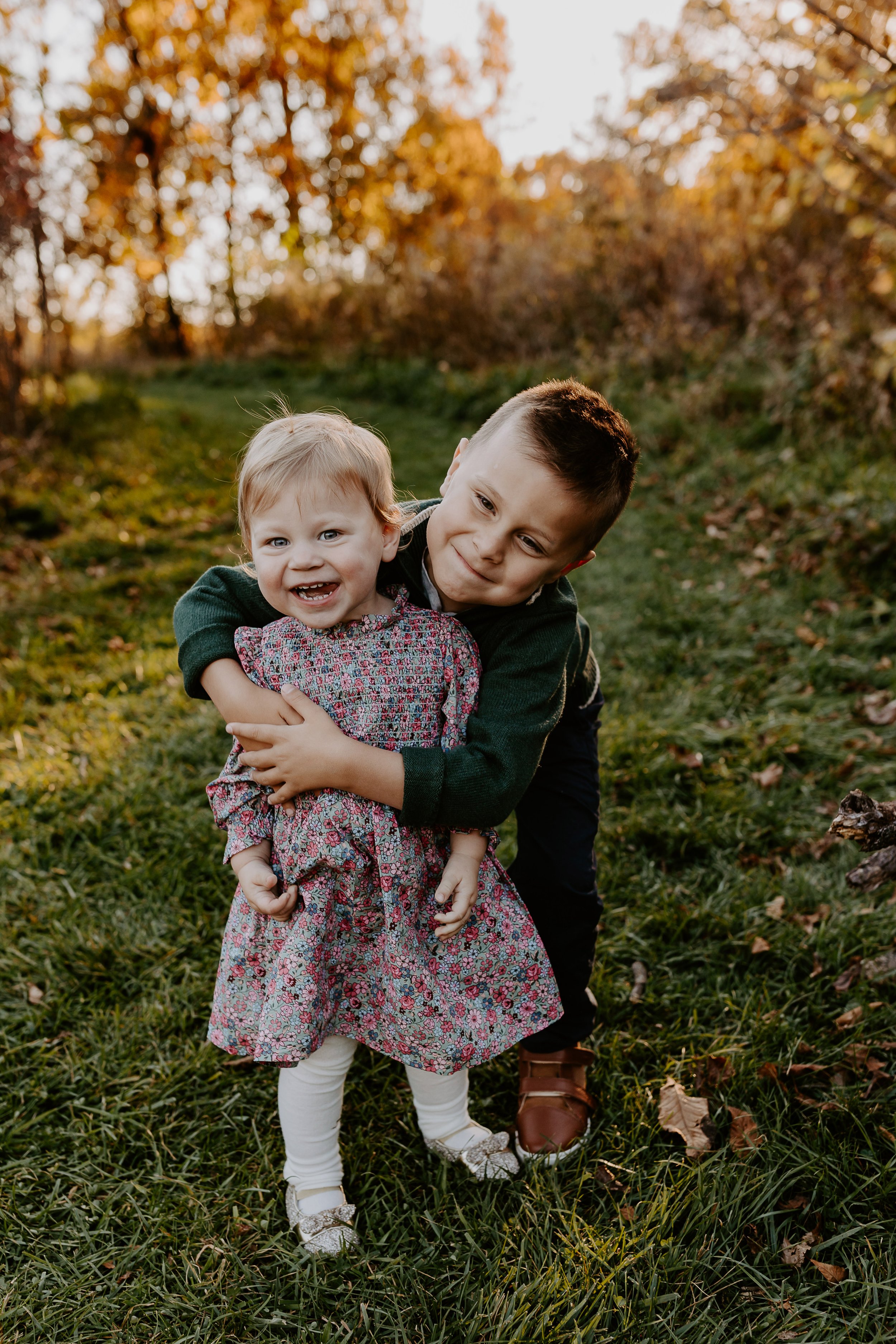 Two children, a boy and a girl, are outdoors on a grassy area during fall, hugging and smiling at the camera, with trees and autumn leaves in the background.