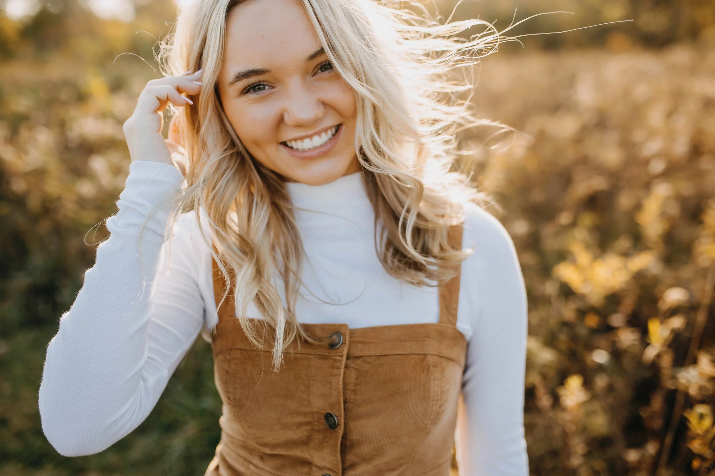 A young woman with blonde hair smiling outdoors in a sunny, autumn setting, wearing a white long-sleeve shirt and a tan corduroy dress.