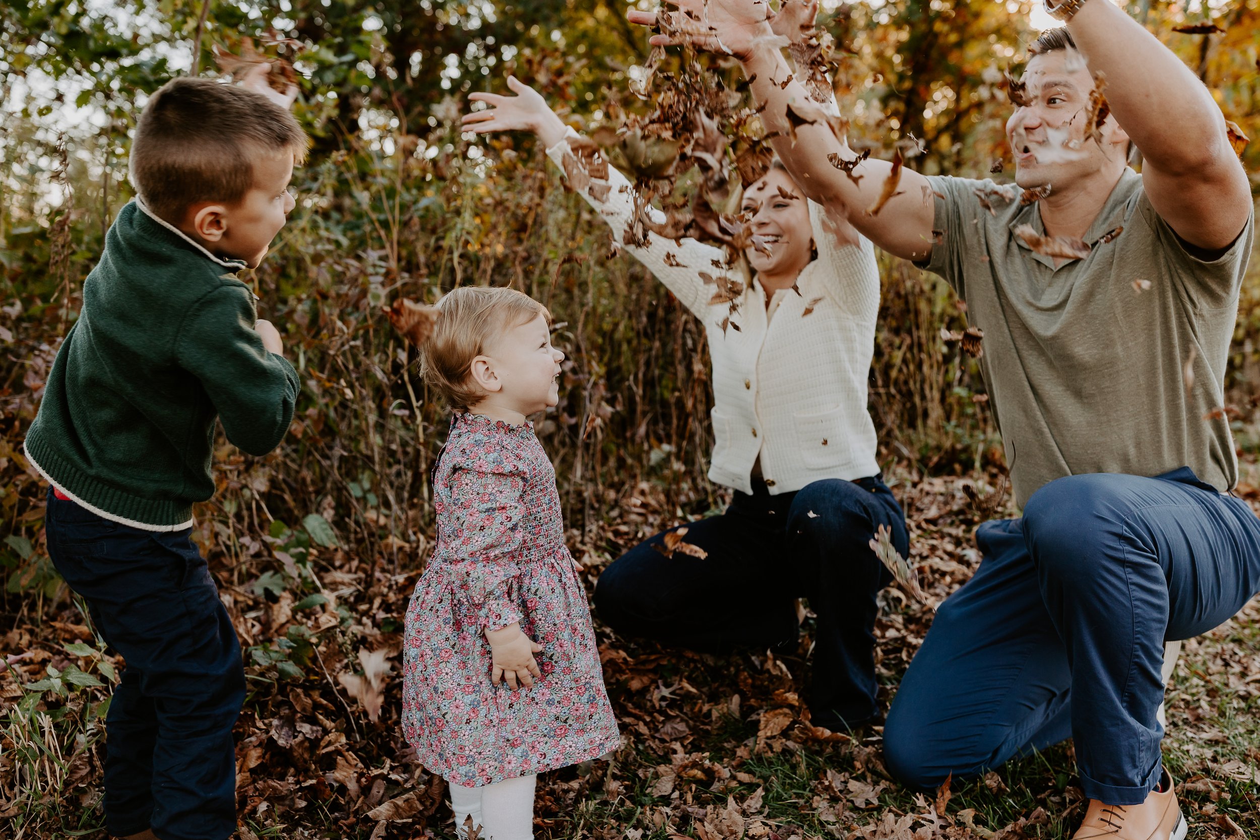 A group of children and two adults playing in a pile of fallen leaves in a wooded area during autumn. The adults are tossing leaves into the air while children watch and enjoy the moment.