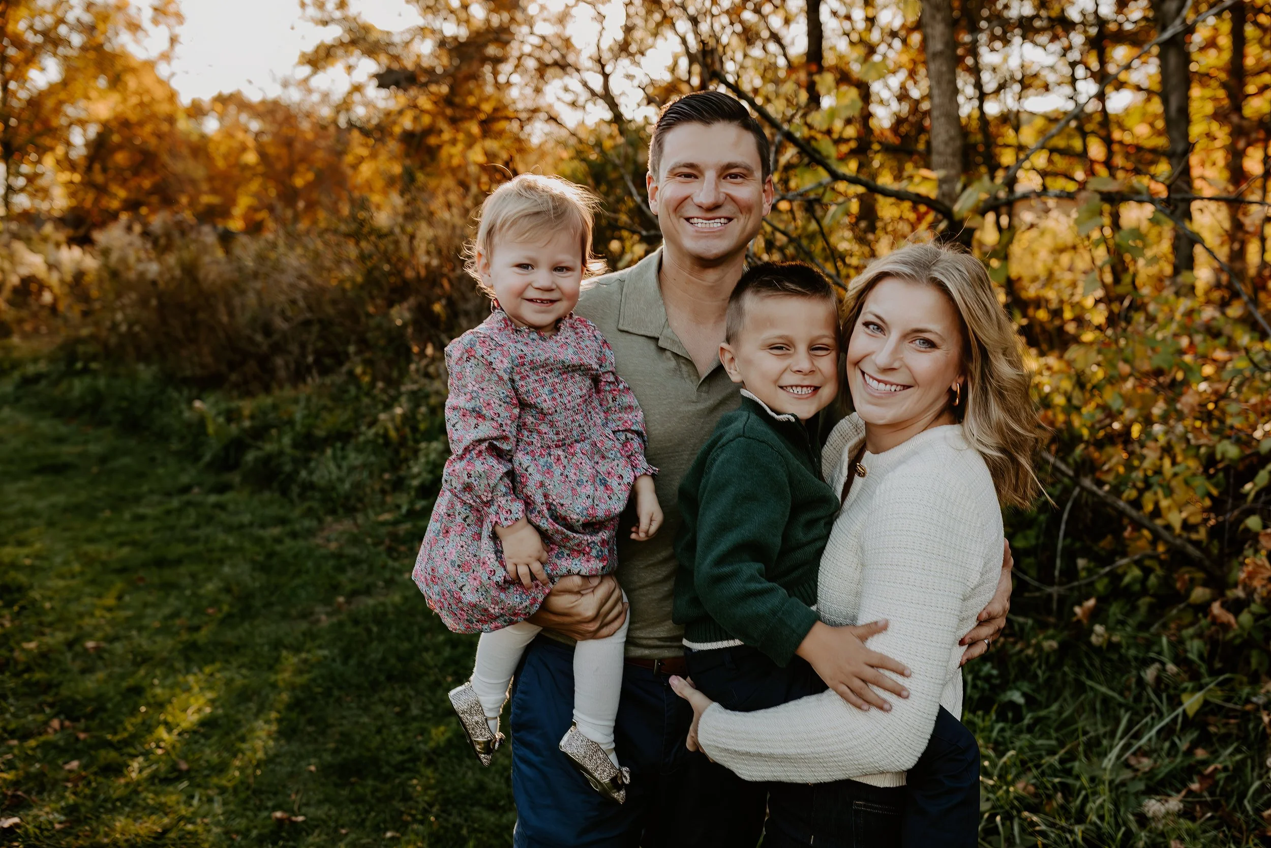 A happy family of four outdoors in autumn, smiling, with autumn leaves in the background.