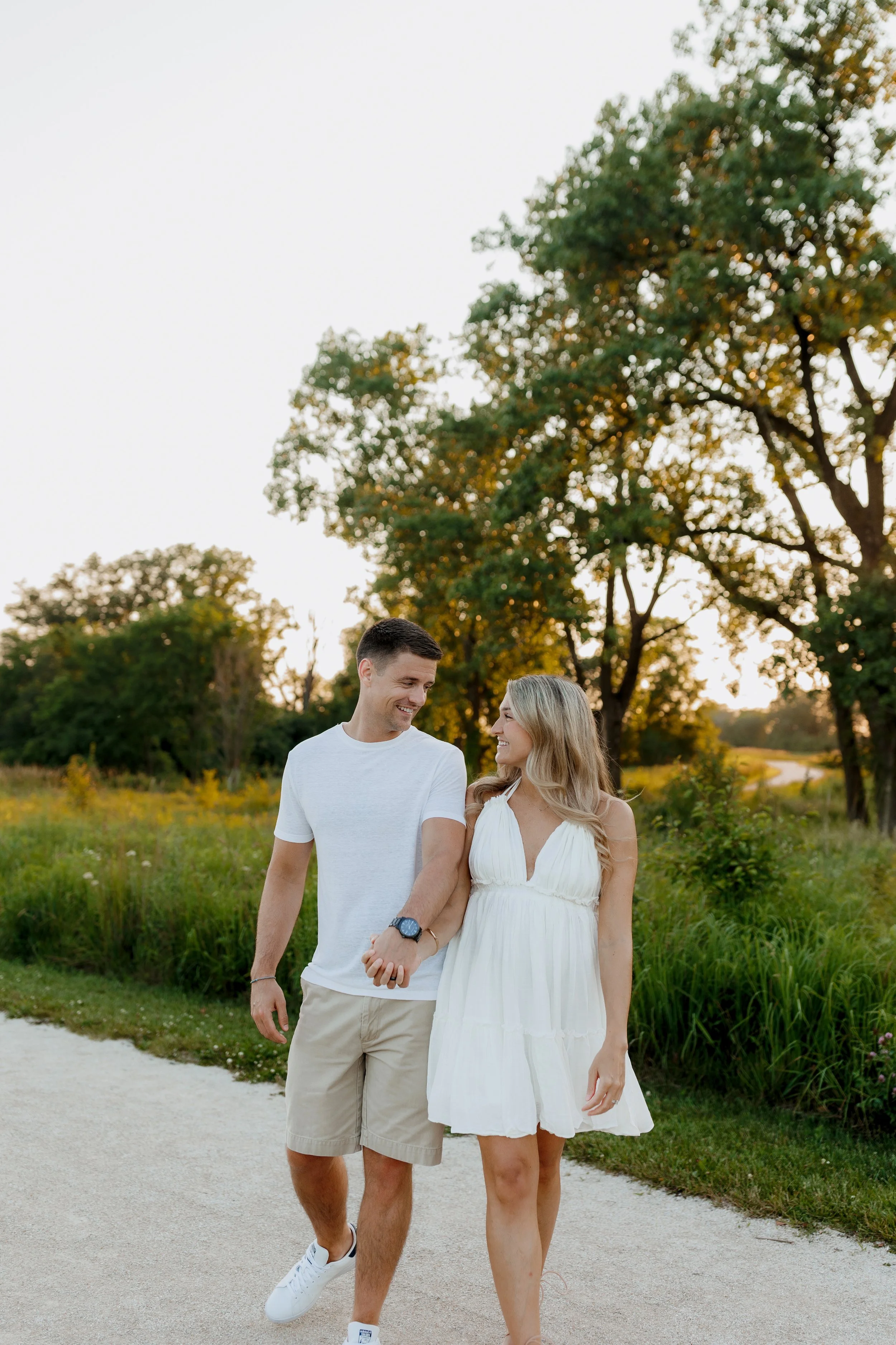 A young couple walking hand in hand outdoors in a park during golden hour. The man is smiling at the woman, who is wearing a white sundress, as they look at each other. The background features green trees and a trail.