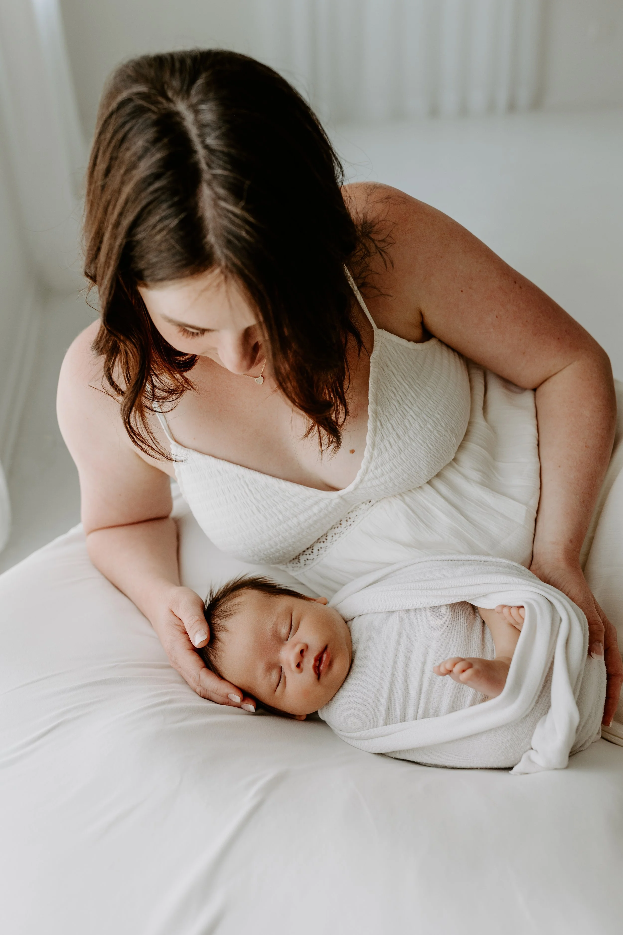 A woman with brown hair, wearing a white dress, is gently holding a sleeping baby with dark hair and closed eyes on a white bed in a softly lit room.