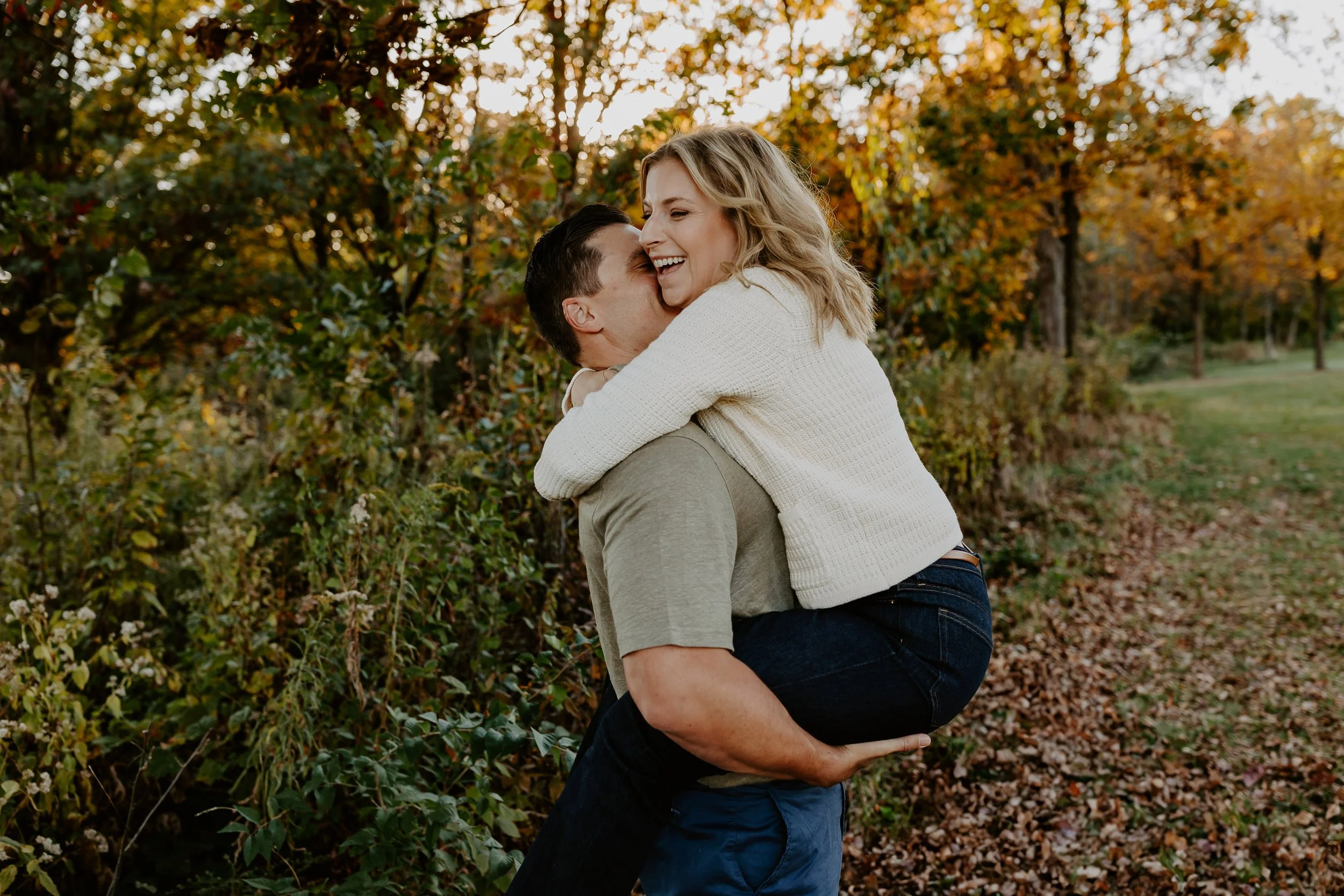 A couple outdoors during fall, with the woman being lifted onto the man's shoulders, both smiling and laughing. The background is filled with autumn trees and fallen leaves.